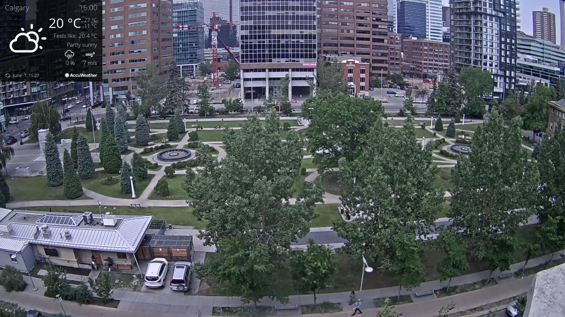 A partly sunny urban park with a fountain and walkways, surrounded by tall buildings under a partly sunny sky.