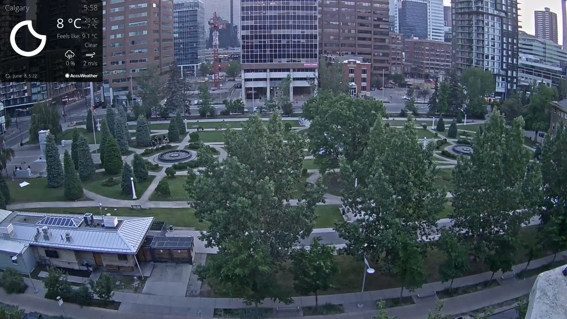 An aerial view of a city park with lush green lawns, paved pathways, and many trees under clear skies.