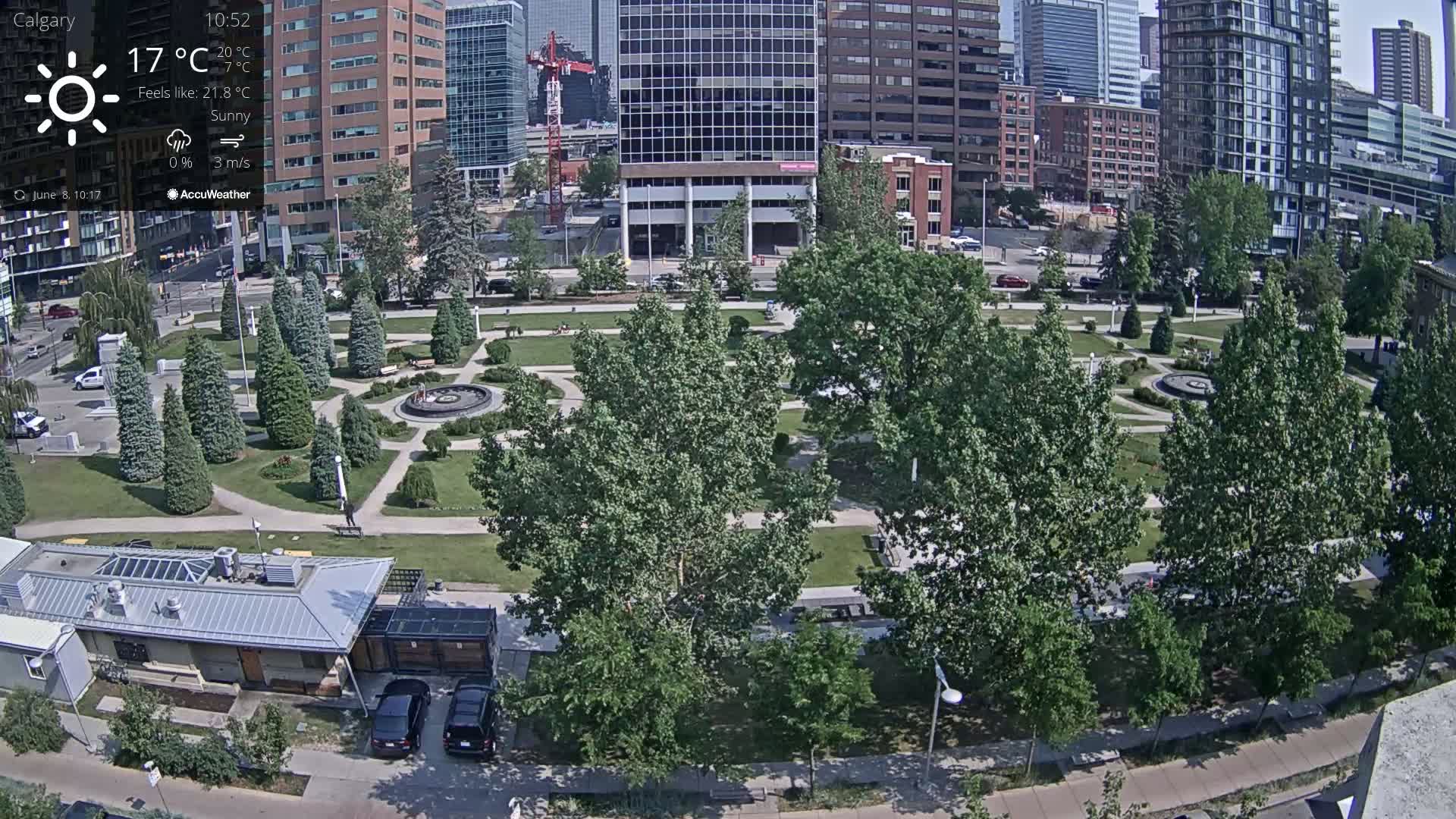 A sunny city park with a central fountain, surrounded by green trees and paved pathways, is situated next to tall buildings under a sunny sky.