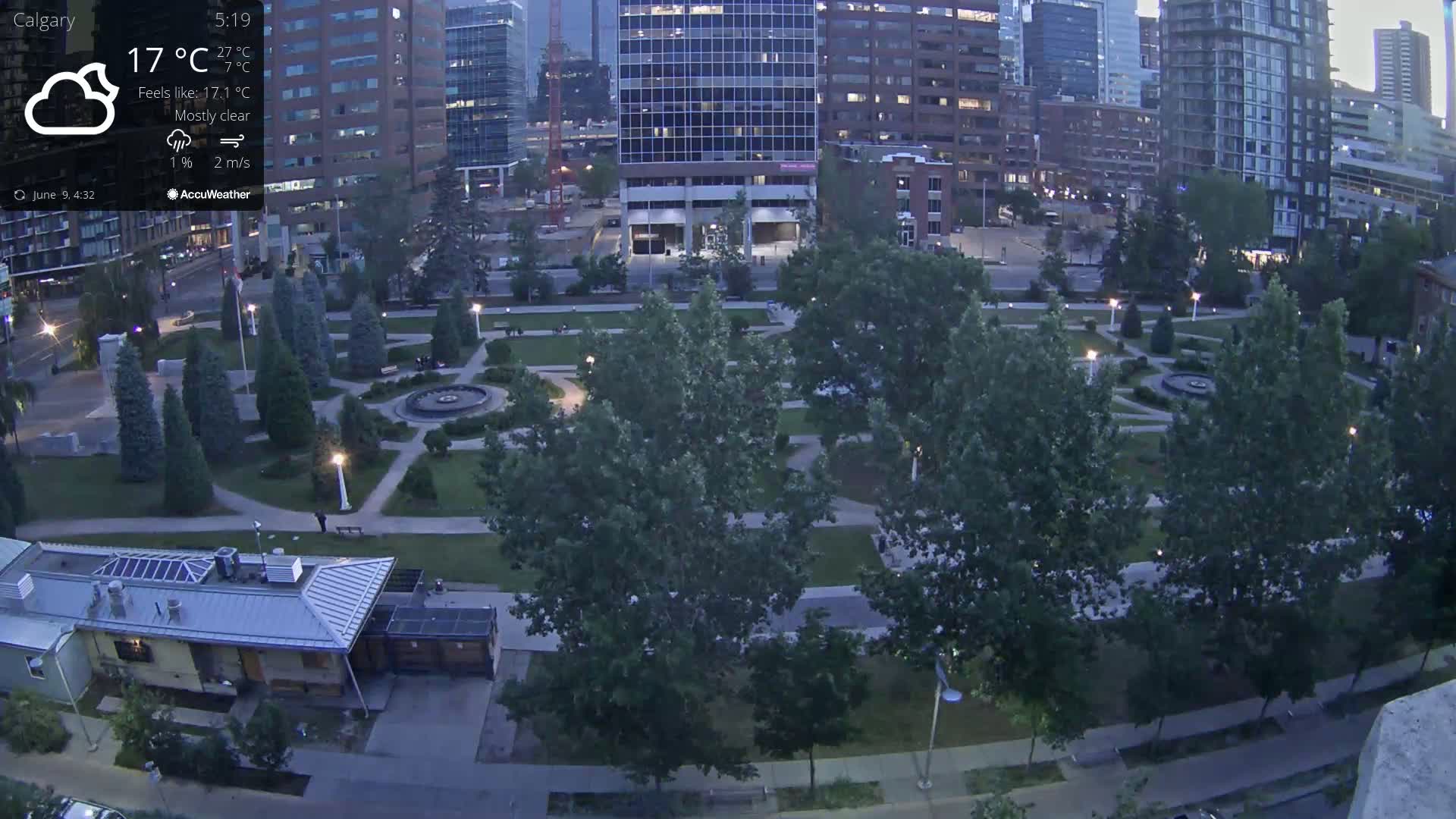 A mostly clear evening scene shows a city park with paved paths, trees, and a central fountain, surrounded by tall buildings under a mostly clear sky.
