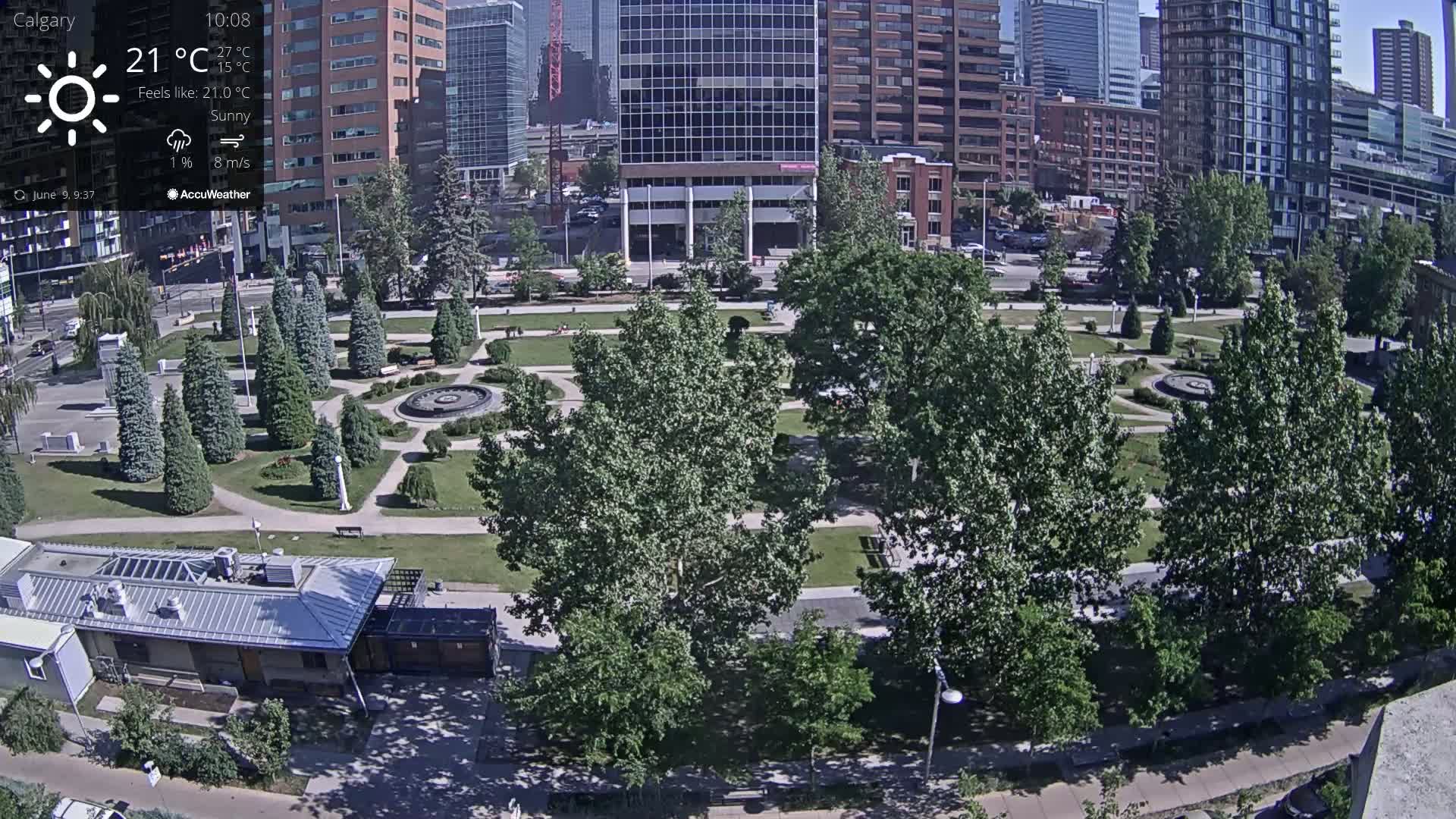 A sunny cityscape with a park containing various trees, shrubs, and a central fountain is visible from an elevated vantage point.