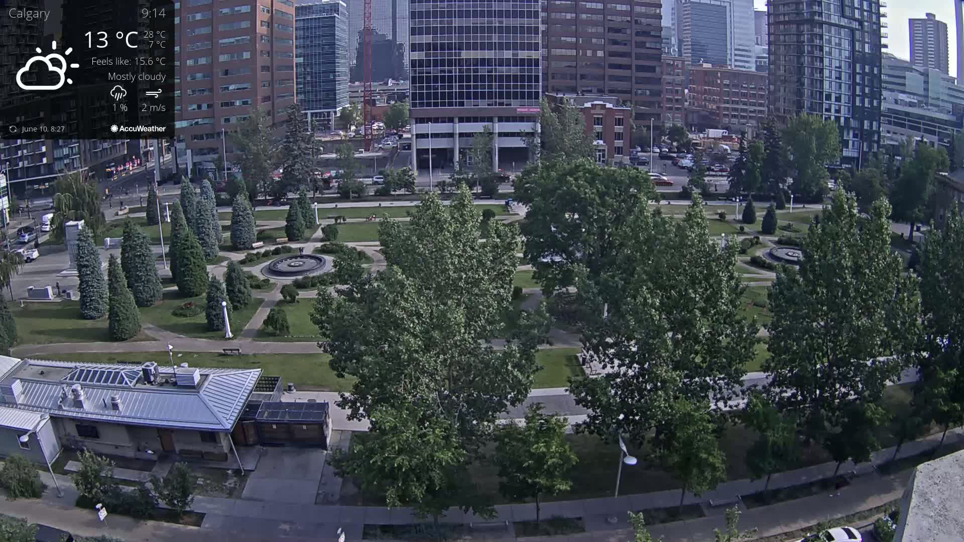 A mostly cloudy day shows an aerial view of a city park with a fountain, surrounded by tall buildings and lush green trees.