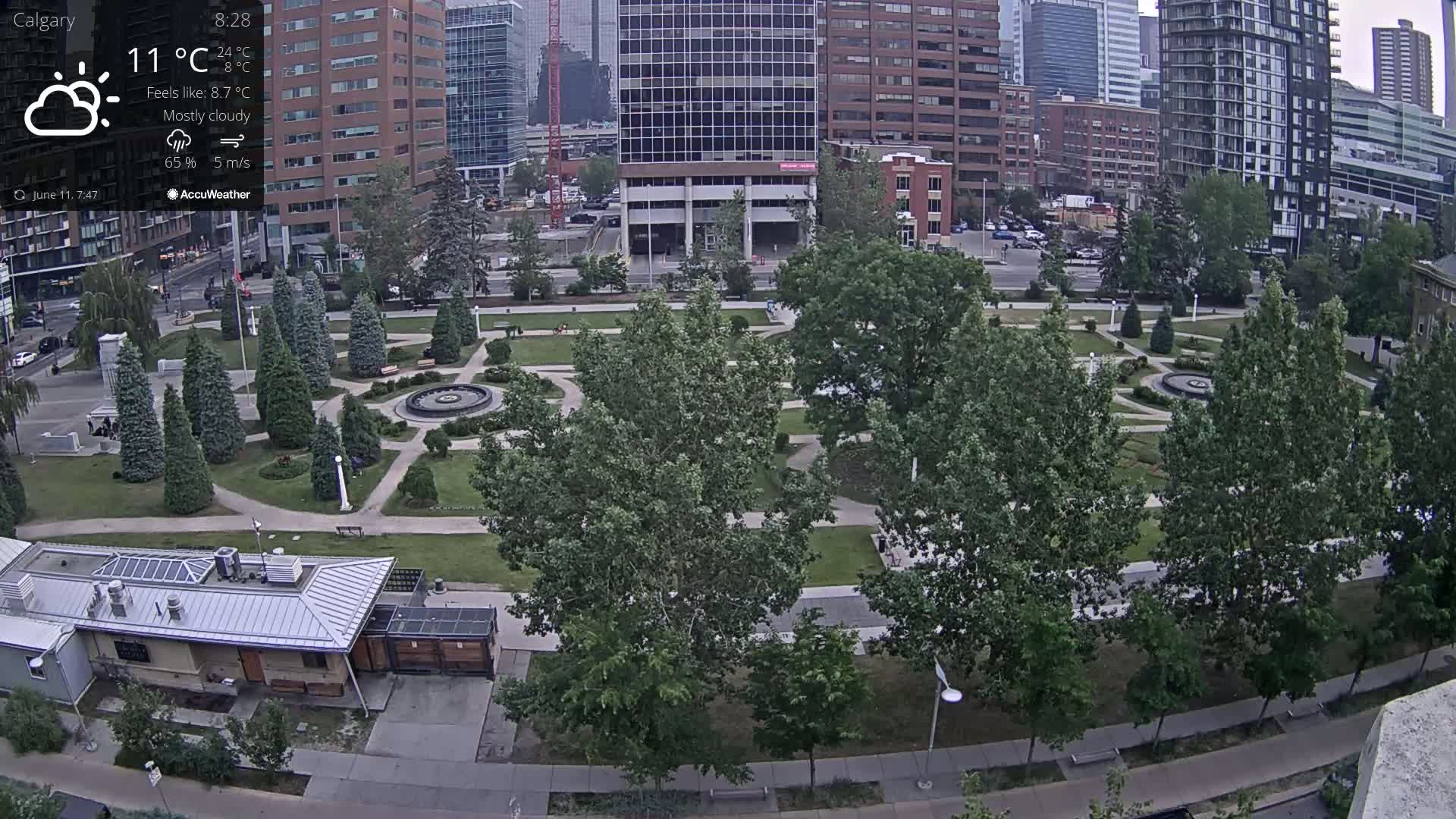 A mostly cloudy city park with a fountain, surrounded by buildings and trees, under a mostly cloudy sky.