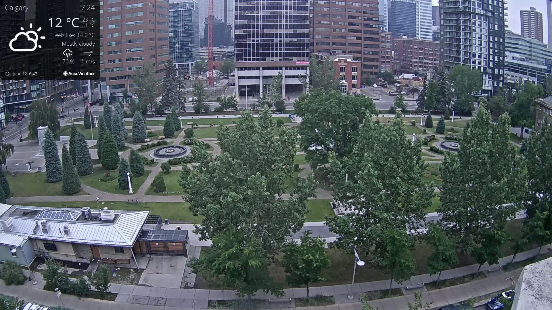 A mostly cloudy city park with a central fountain is surrounded by tall buildings under a mostly cloudy sky.
