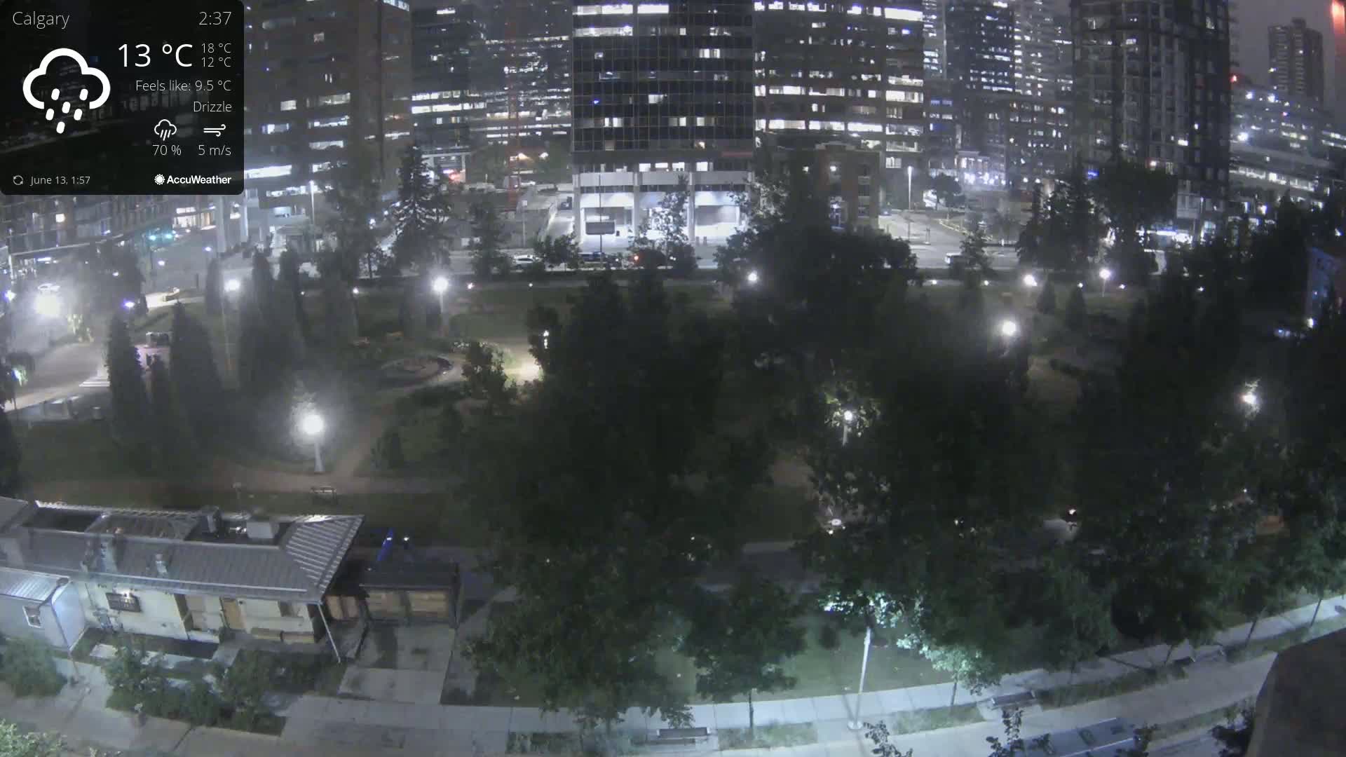 A nighttime view of a park surrounded by city buildings under a drizzle.