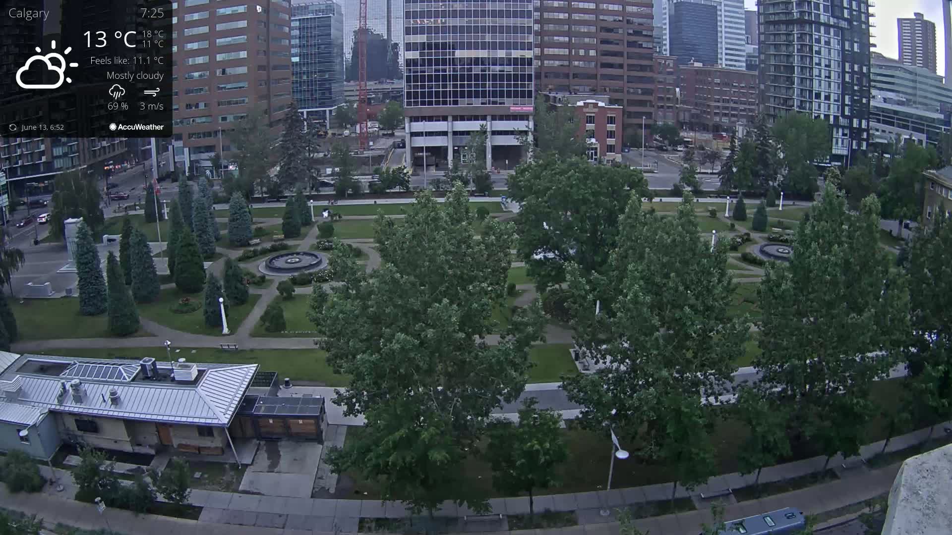 A mostly cloudy city park with a fountain, surrounded by tall buildings and lush green trees under a partly sunny sky.