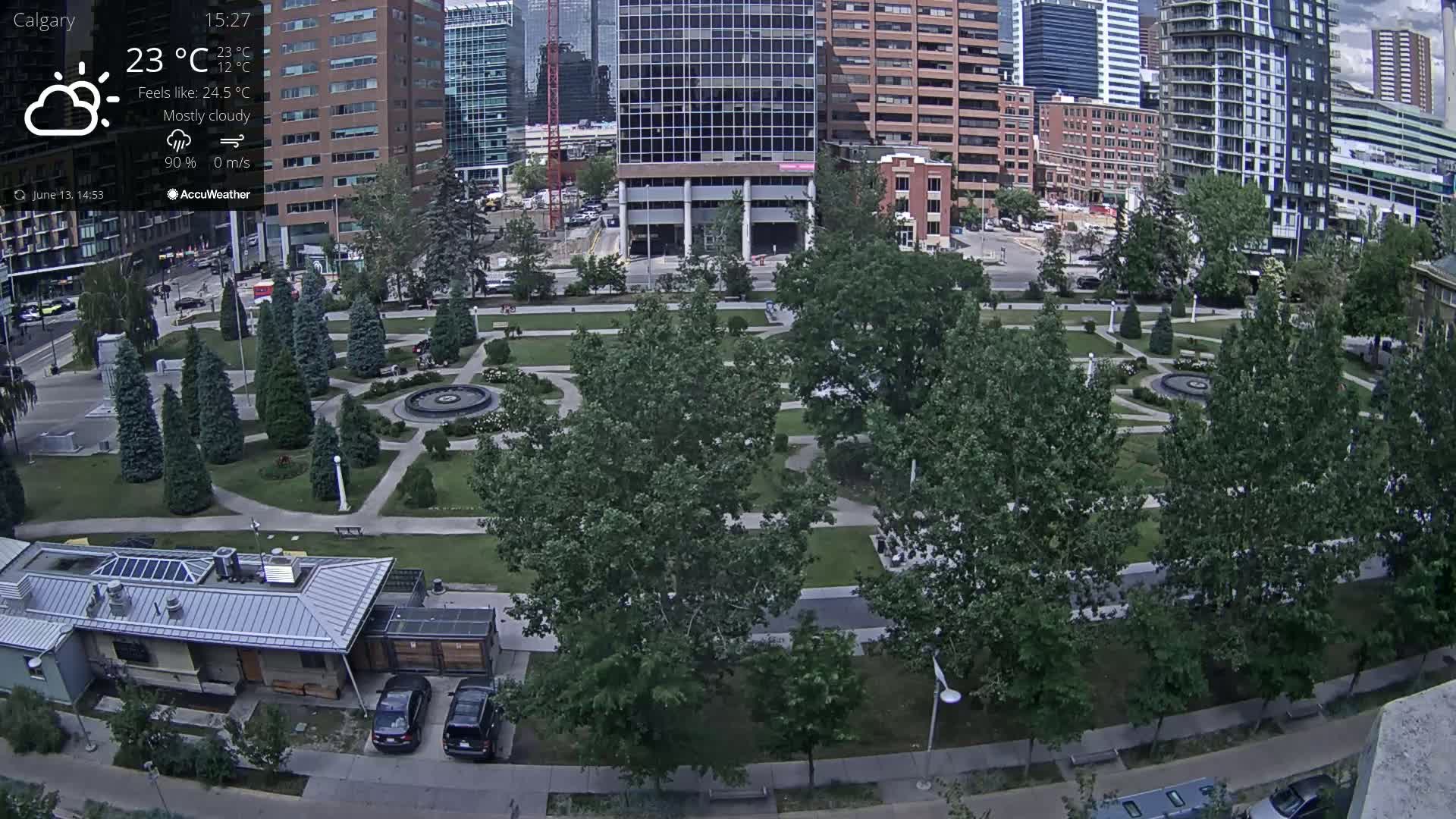A mostly cloudy city park with a central fountain is visible from an elevated viewpoint, surrounded by tall buildings and featuring several parked cars.