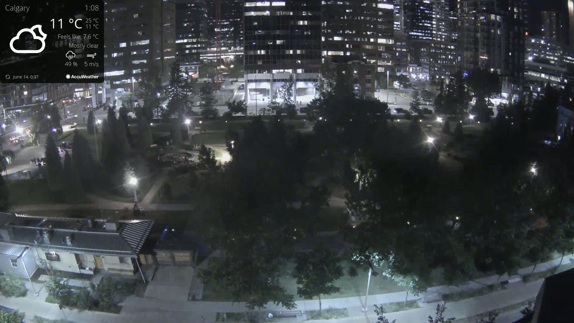 A mostly cloudy city park with a central fountain is visible from an elevated viewpoint, surrounded by tall buildings and featuring several parked cars.