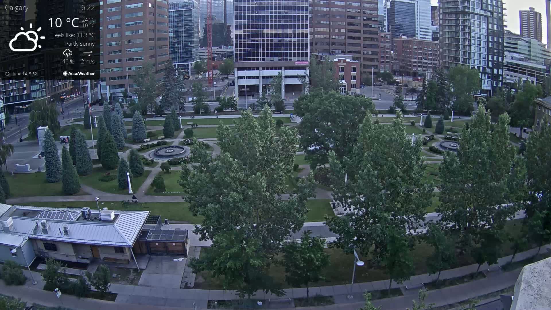 A partly sunny view of a city park with a fountain, surrounded by buildings and lush green trees.