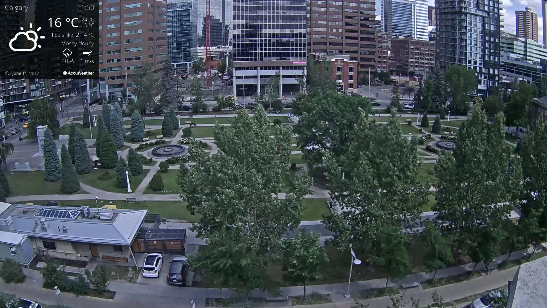 An aerial view of a city park on a mostly cloudy day, showing green lawns, trees, a fountain, and several buildings in the background.