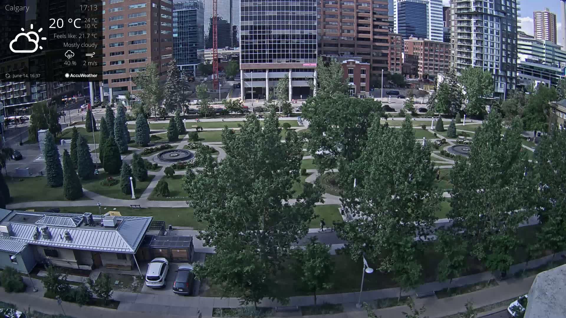 An aerial view of a city park on a mostly cloudy day, showing green lawns, trees, a fountain, and several buildings in the background.