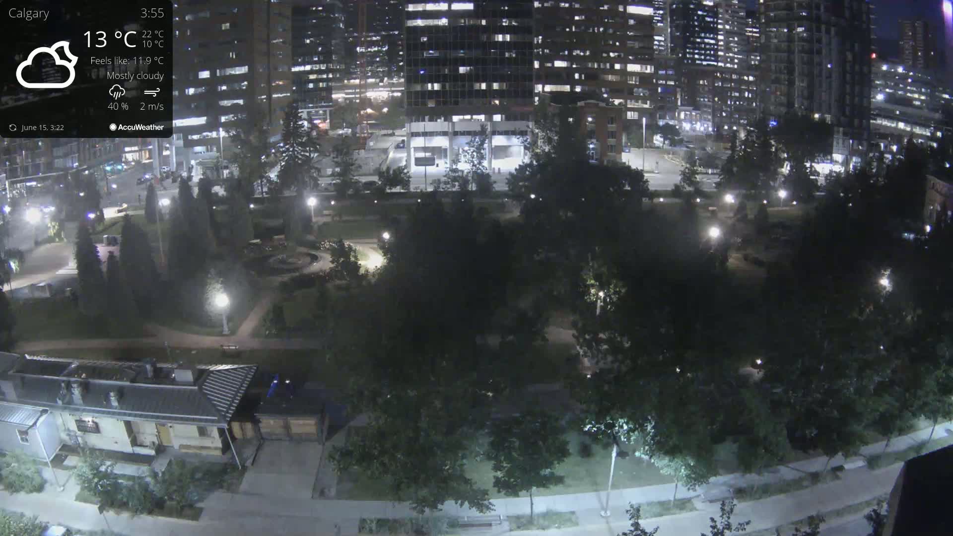 A mostly cloudy night scene shows a park with trees and pathways, bordered by city buildings and lit by streetlights.