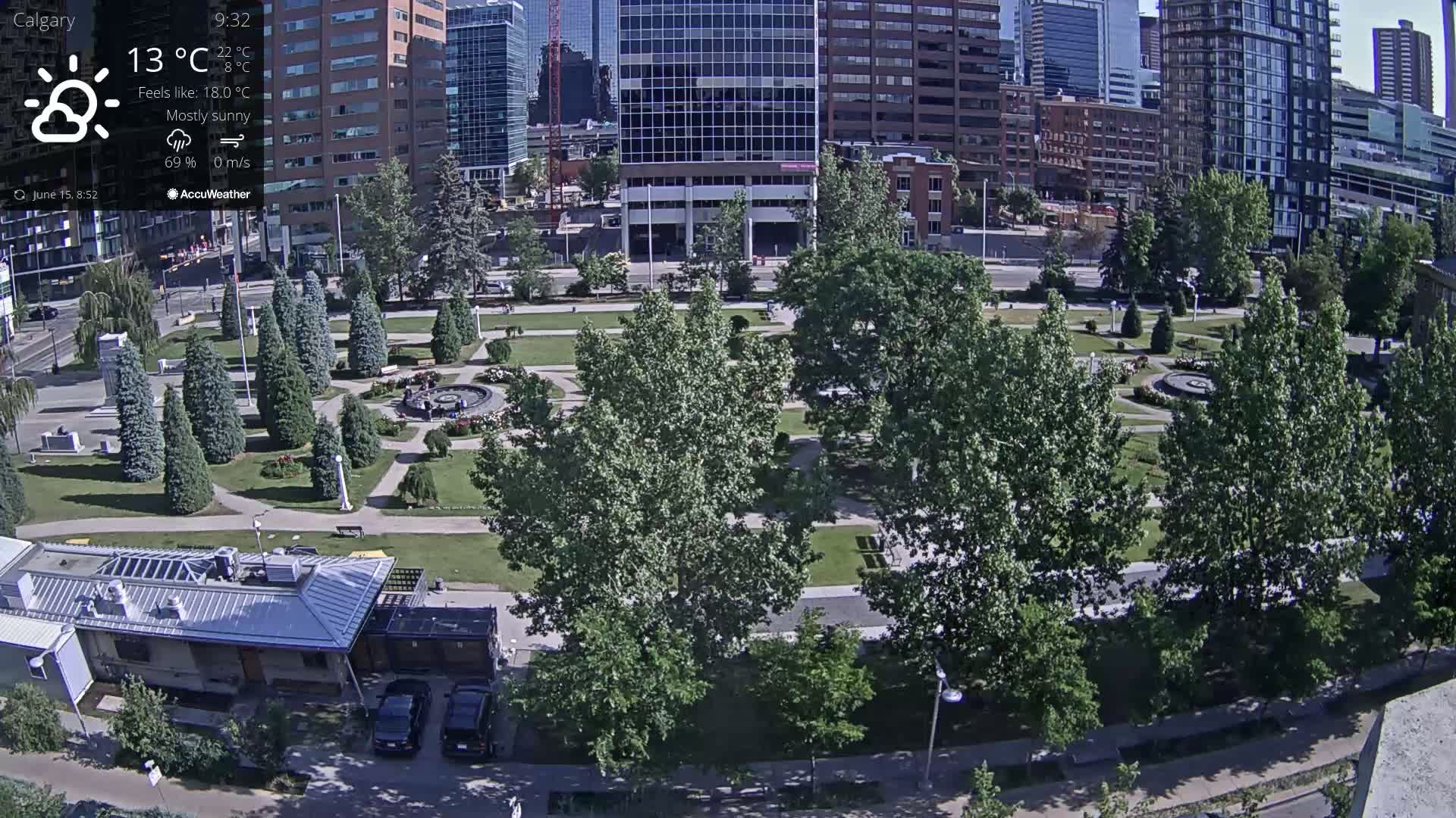 A mostly sunny city park with a fountain, surrounded by tall buildings and lush green trees.