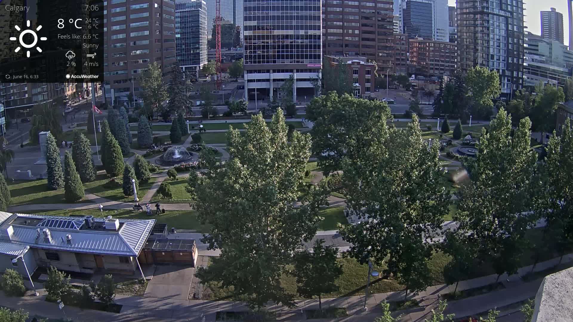 An aerial view of a city park with a water fountain, surrounded by tall buildings under sunny conditions.