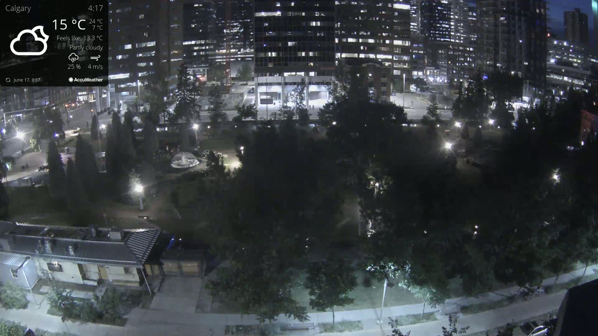 An aerial nighttime view of a city park with partly cloudy conditions, showing a grassy area surrounded by trees and buildings with lights illuminating the scene.