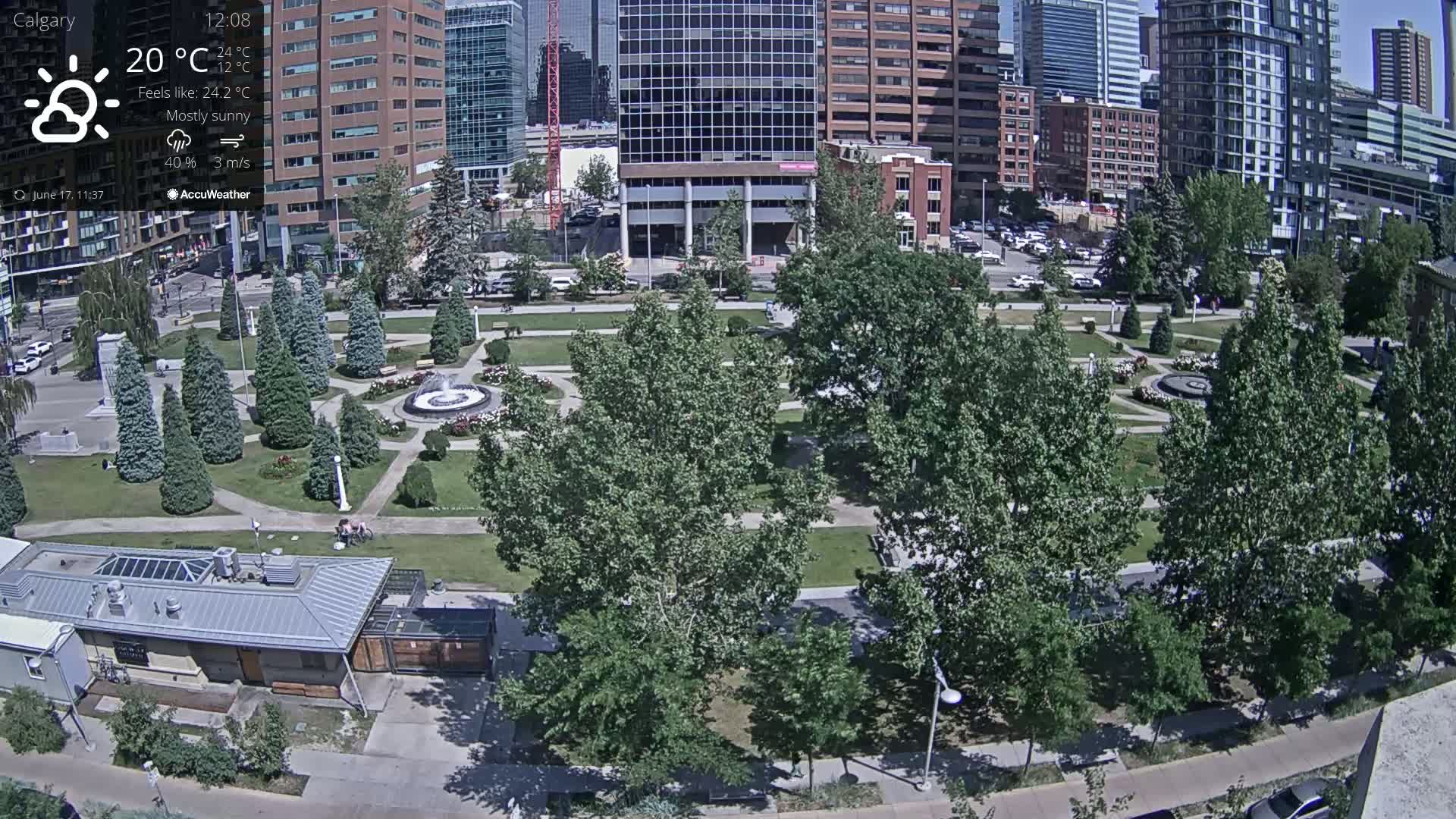 A mostly sunny city park with a fountain, surrounded by tall buildings, under a mostly sunny sky with a temperature of 20°C.
