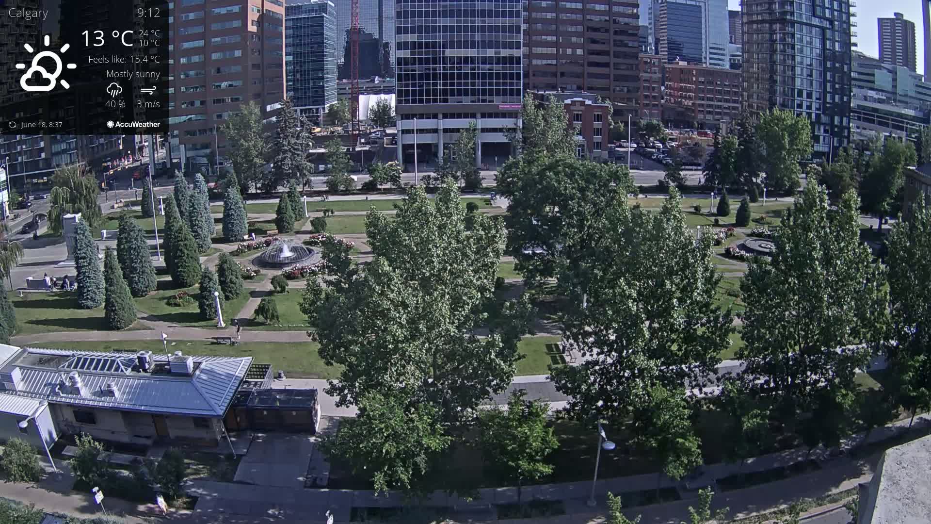 A mostly sunny city park with a fountain is surrounded by tall buildings and trees.