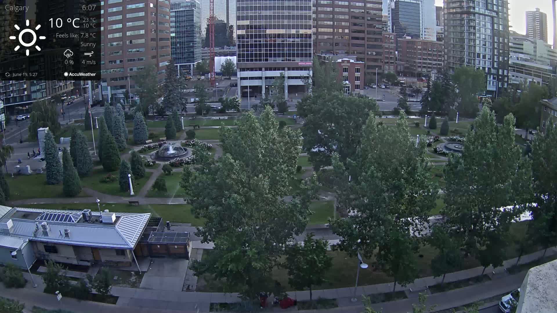 A sunny city park with a fountain is surrounded by tall buildings and trees on a partly cloudy day.