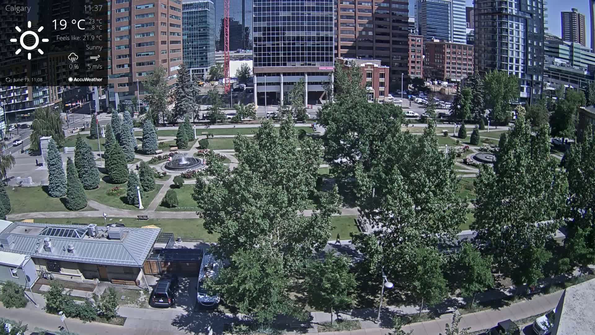 A sunny city park with a fountain, surrounded by tall buildings and lush green trees under a sunny sky.