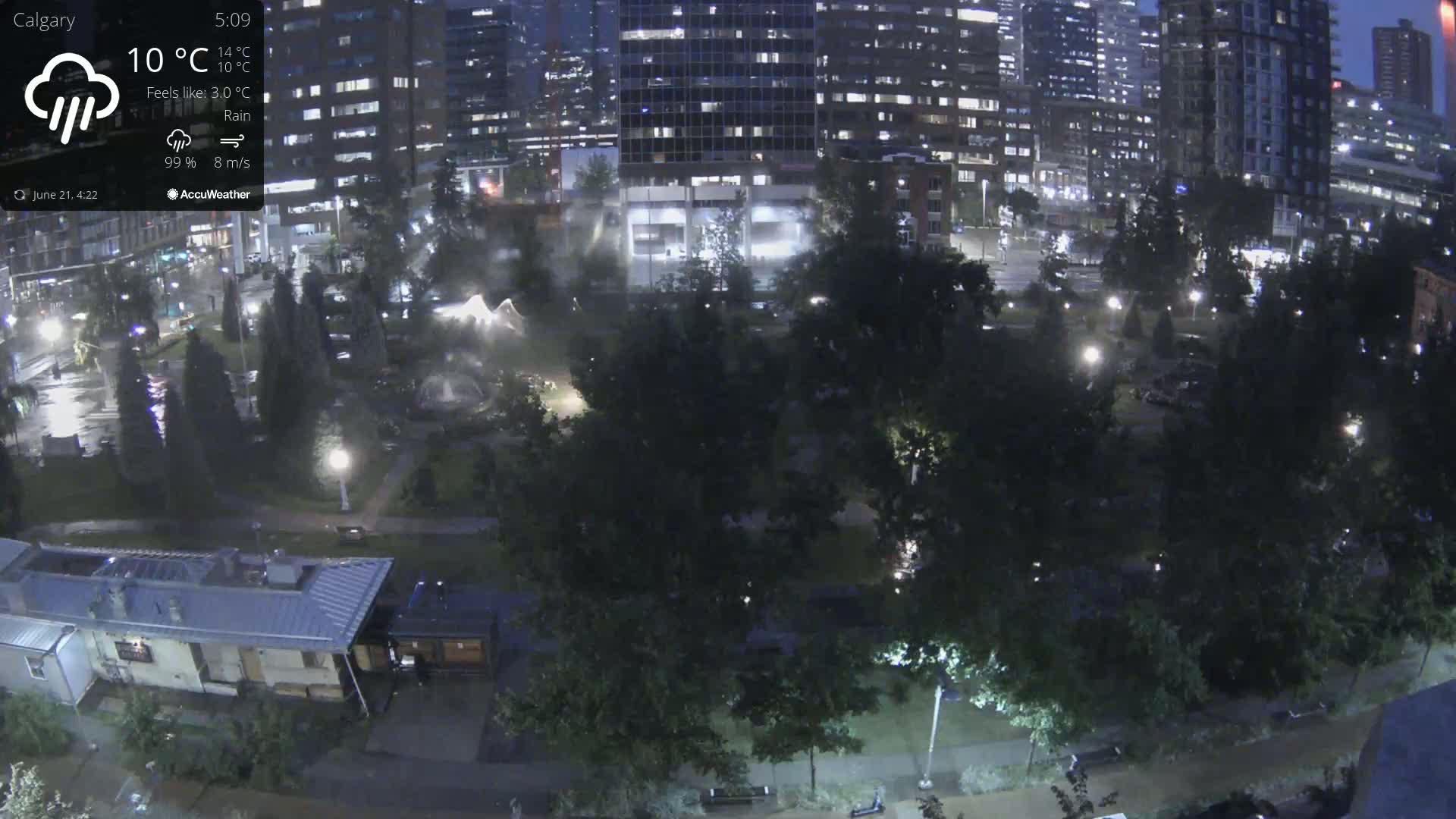 A nighttime view of a city park with tall buildings in the background during a rainy night.