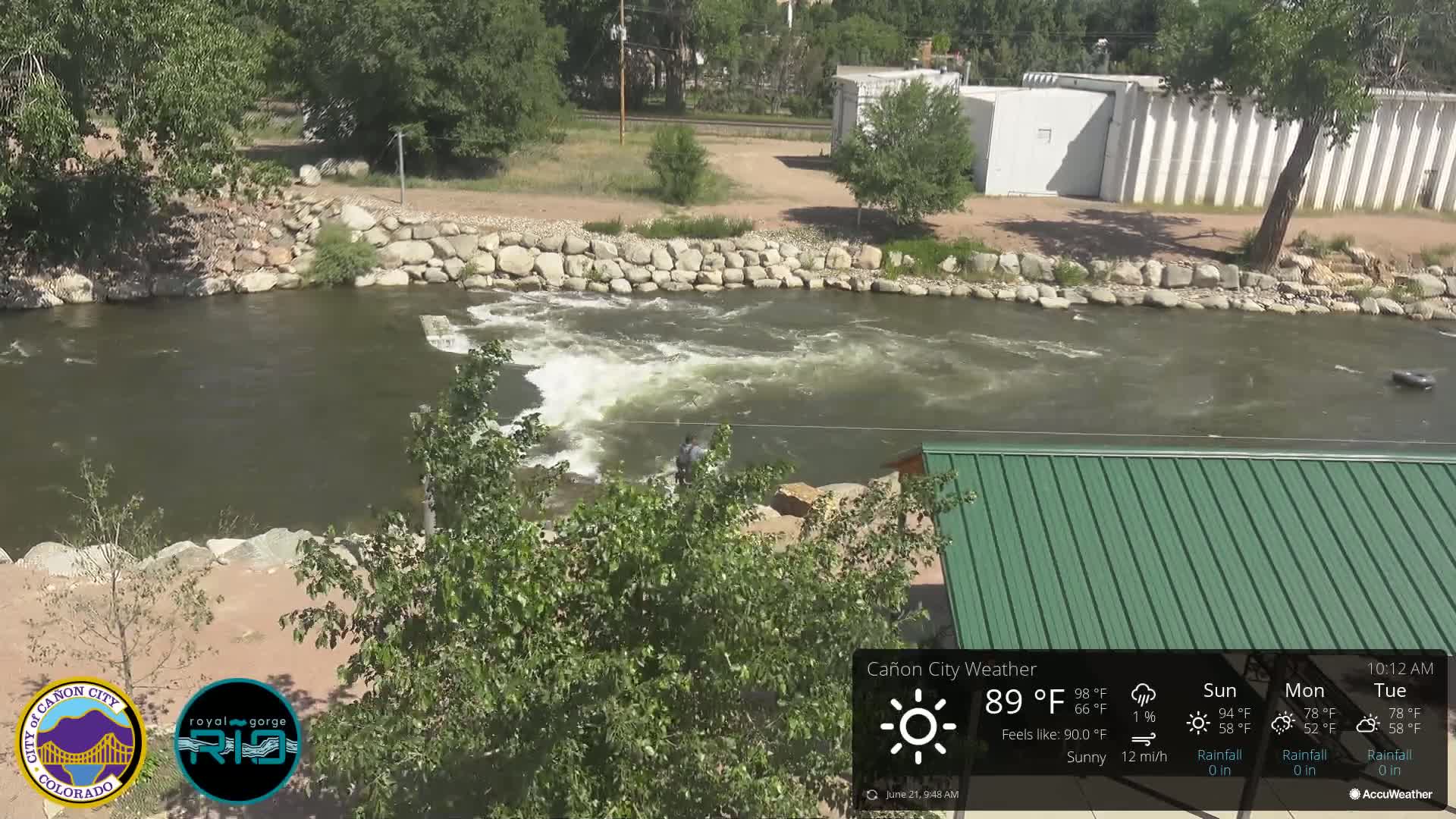 A person stands near a rocky riverbank where a river flows rapidly over a small dam under sunny conditions.