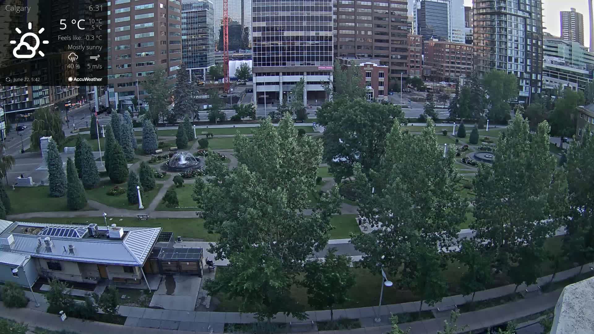 An aerial view shows a city park with a fountain and many green trees under mostly sunny skies.
