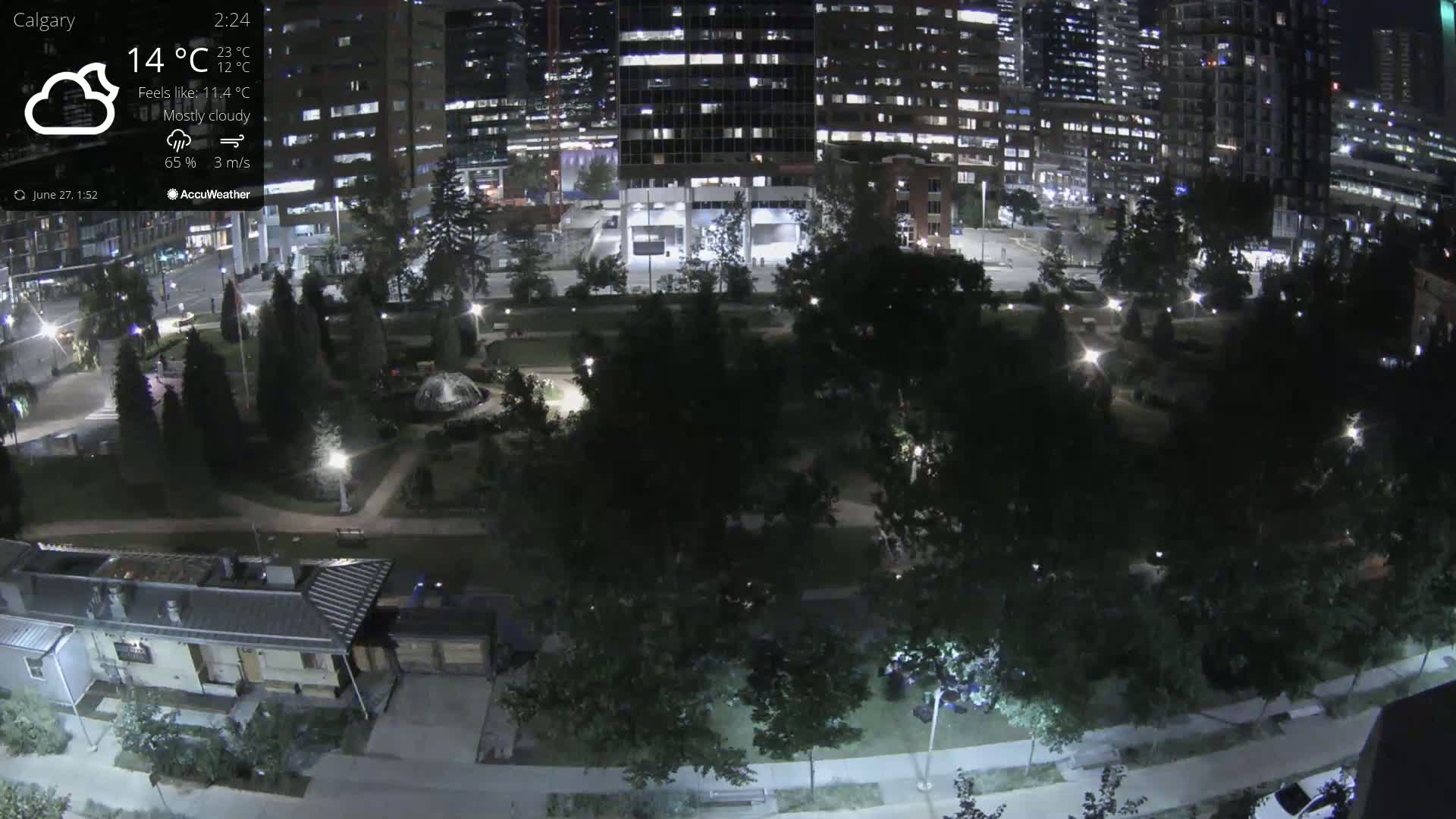 A mostly cloudy night scene shows a park with walkways and trees, bordered by illuminated city buildings.
