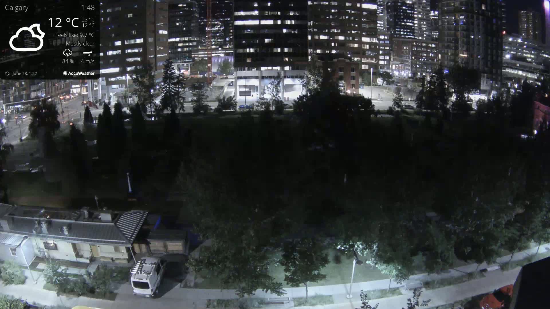 A nighttime view of a city park, mostly dark with some streetlights illuminating paths and trees, adjacent to brightly lit tall buildings under mostly clear skies.
