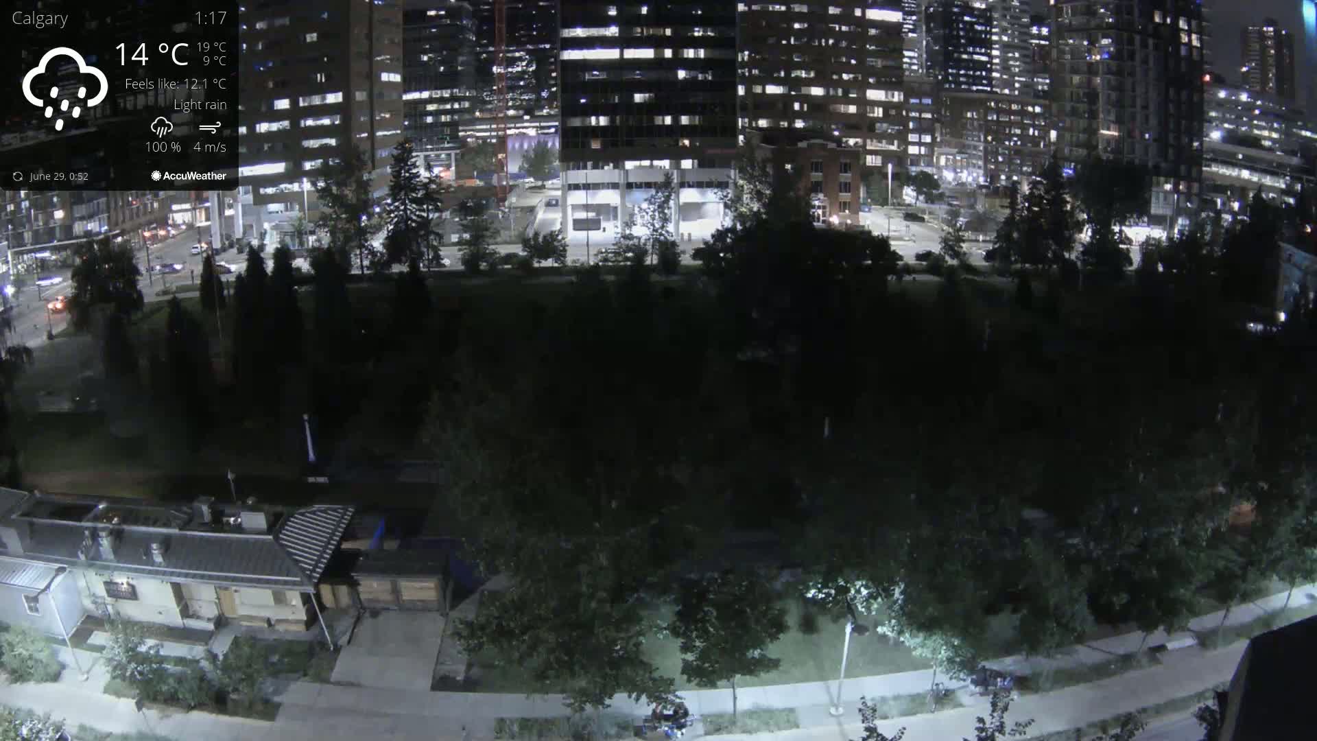 A nighttime view of a park with light rain, illuminated by streetlights, adjacent to brightly lit city buildings.