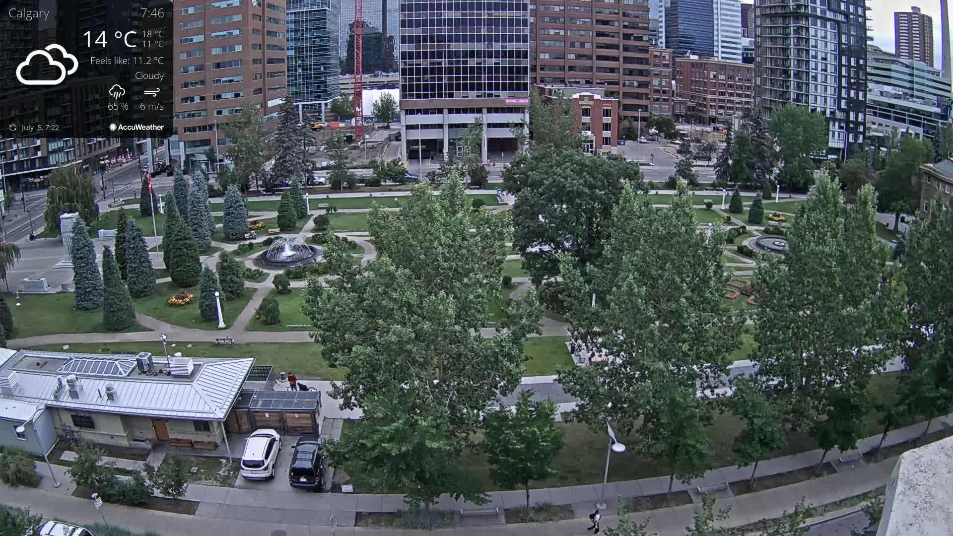 A city park with a fountain in the center is surrounded by lush green trees and paved walkways, under cloudy skies.