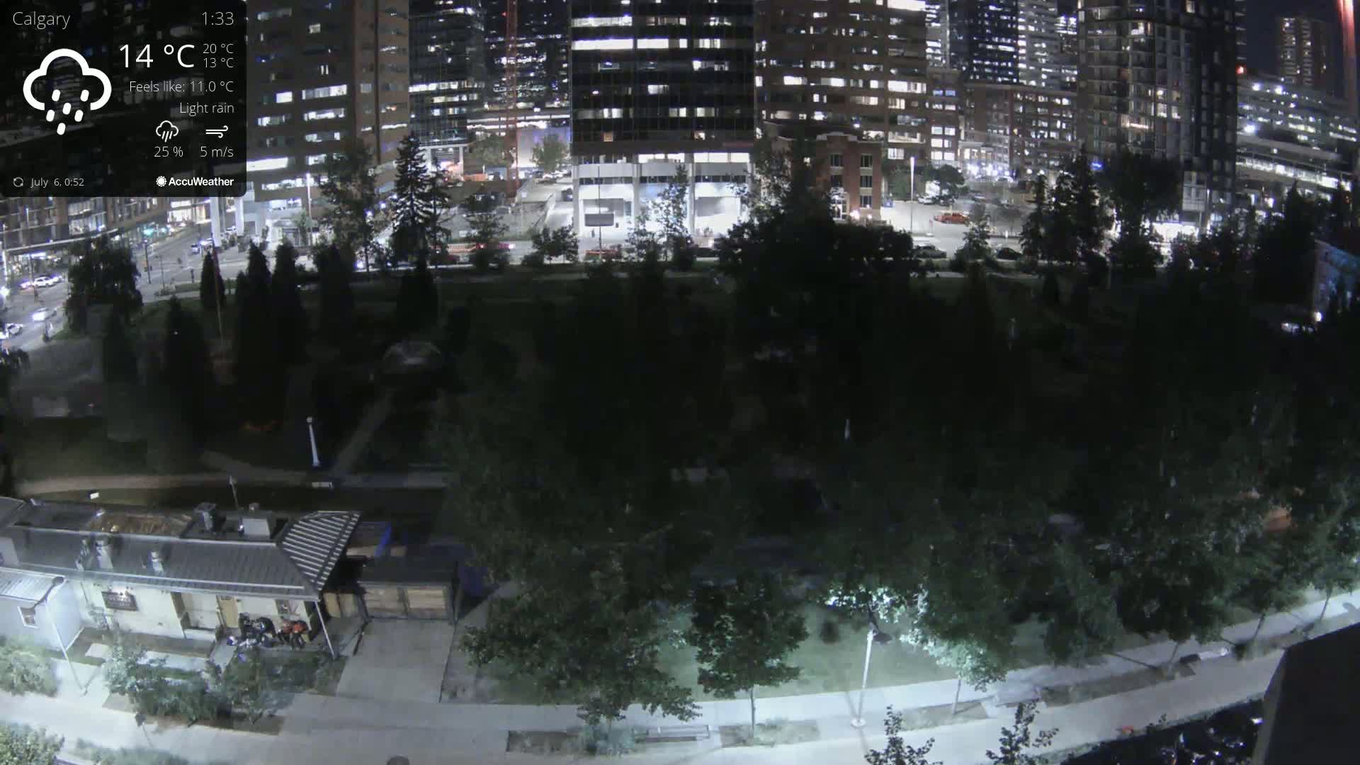 A nighttime view of a city park bordered by illuminated buildings under light rain.