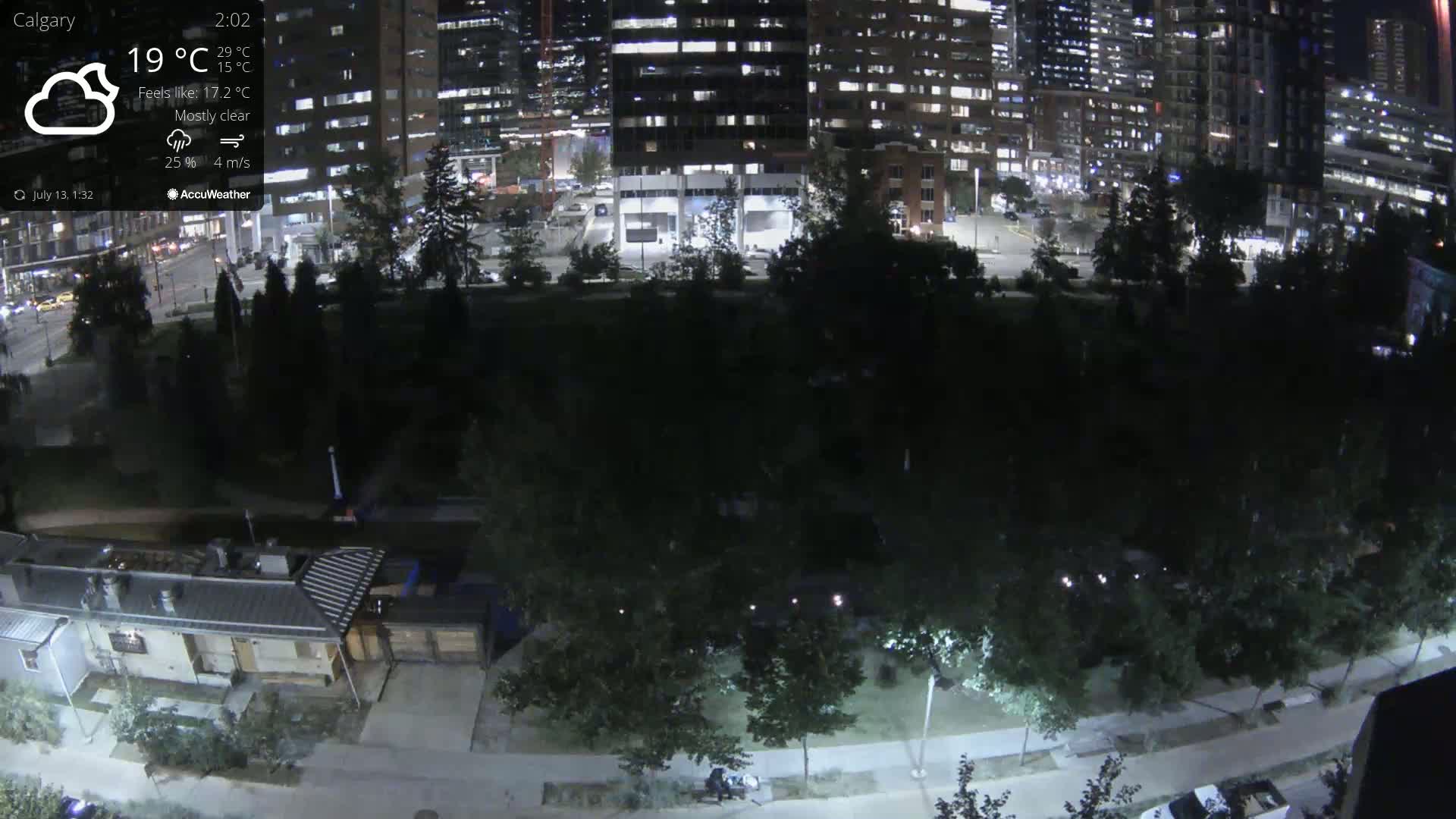 A nighttime, mostly clear view of a city park with surrounding illuminated buildings and a few cars in a parking lot.