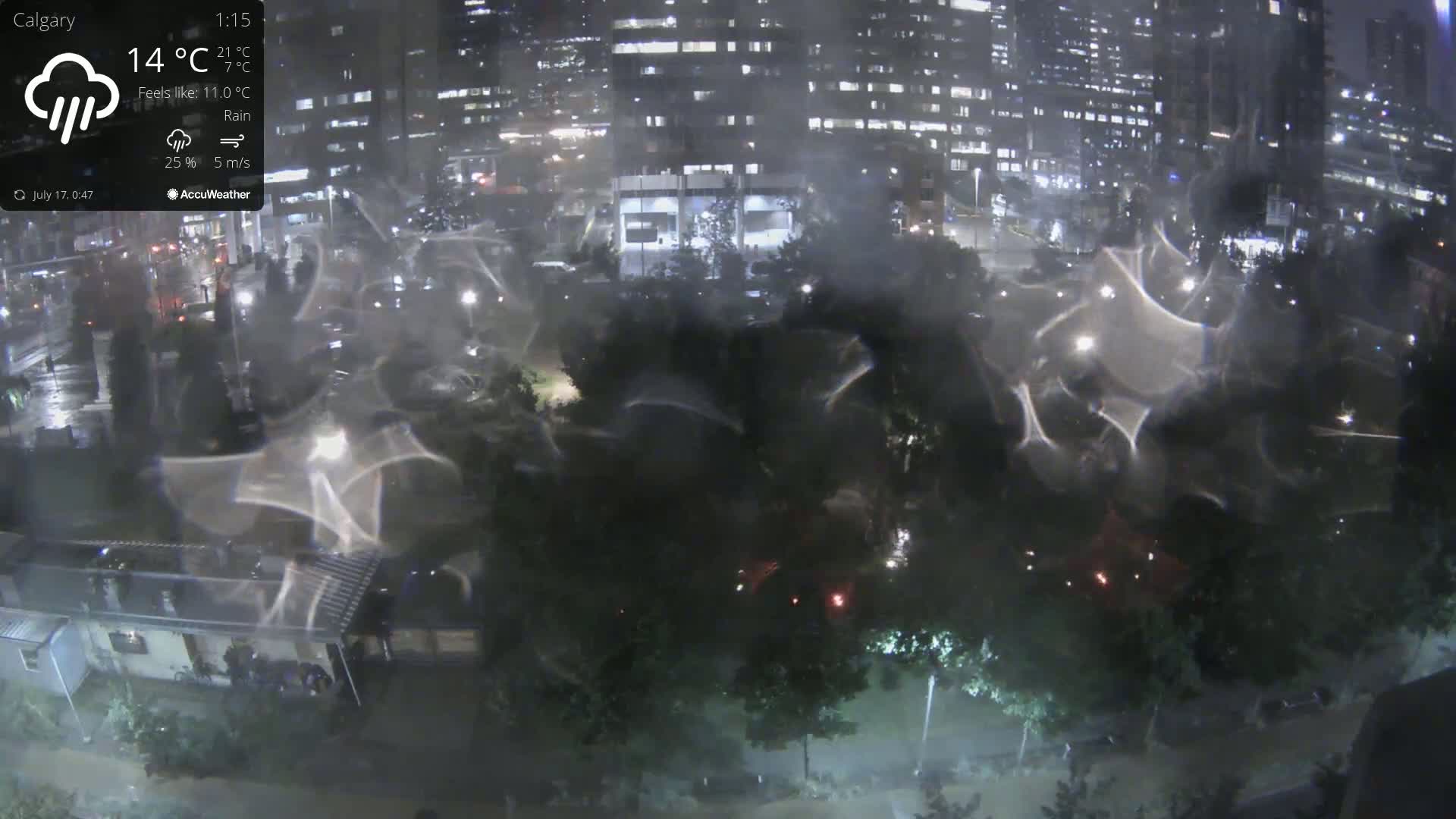 A nighttime view of a city park and surrounding buildings during a rainy period, showing rain-streaked lights and reflections on a wet surface.