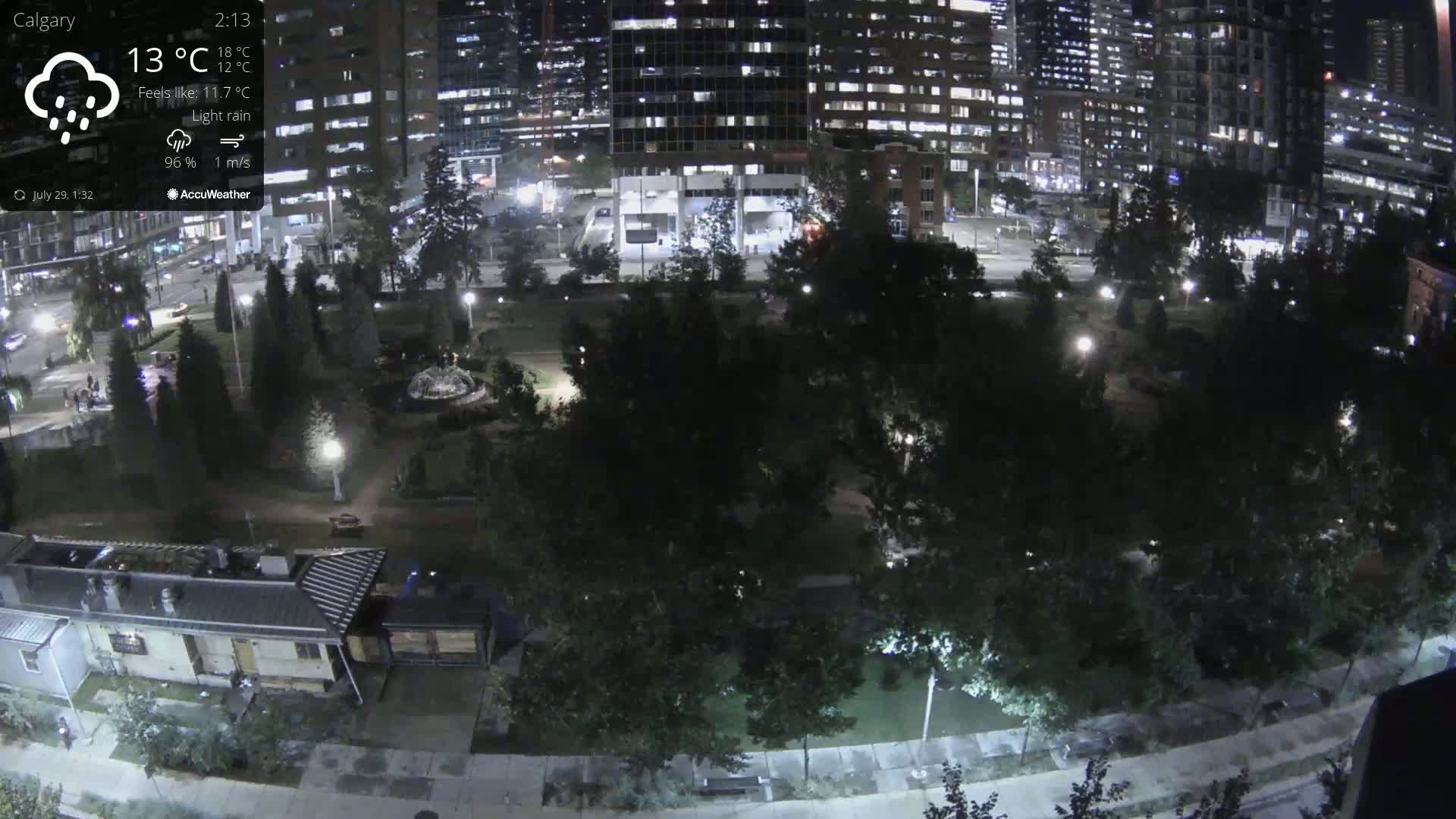 A nighttime, lightly raining view of a city park surrounded by illuminated buildings and trees.