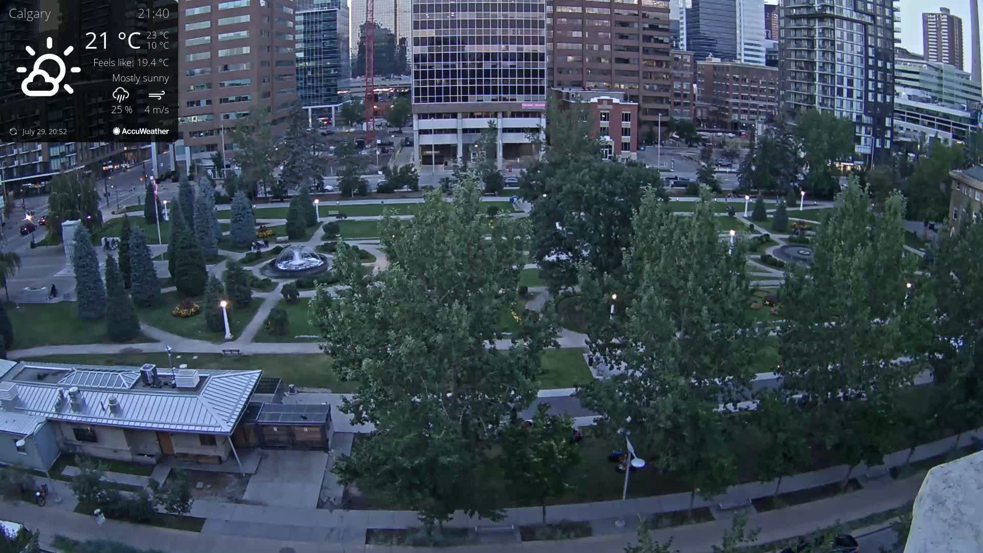 A nighttime, lightly raining view of a city park surrounded by illuminated buildings and trees.
