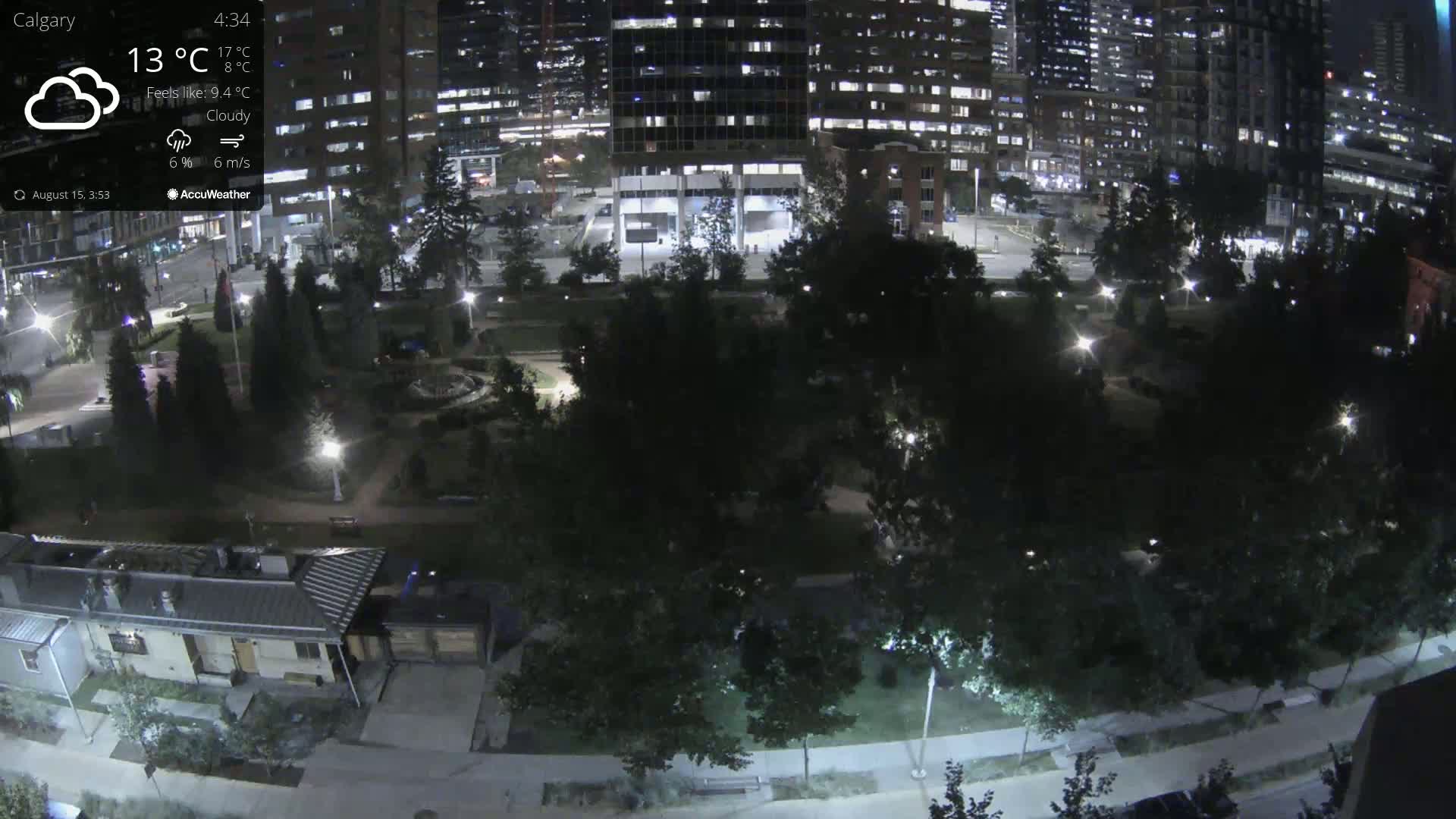 A nighttime, lightly raining view of a city park surrounded by illuminated buildings and trees.