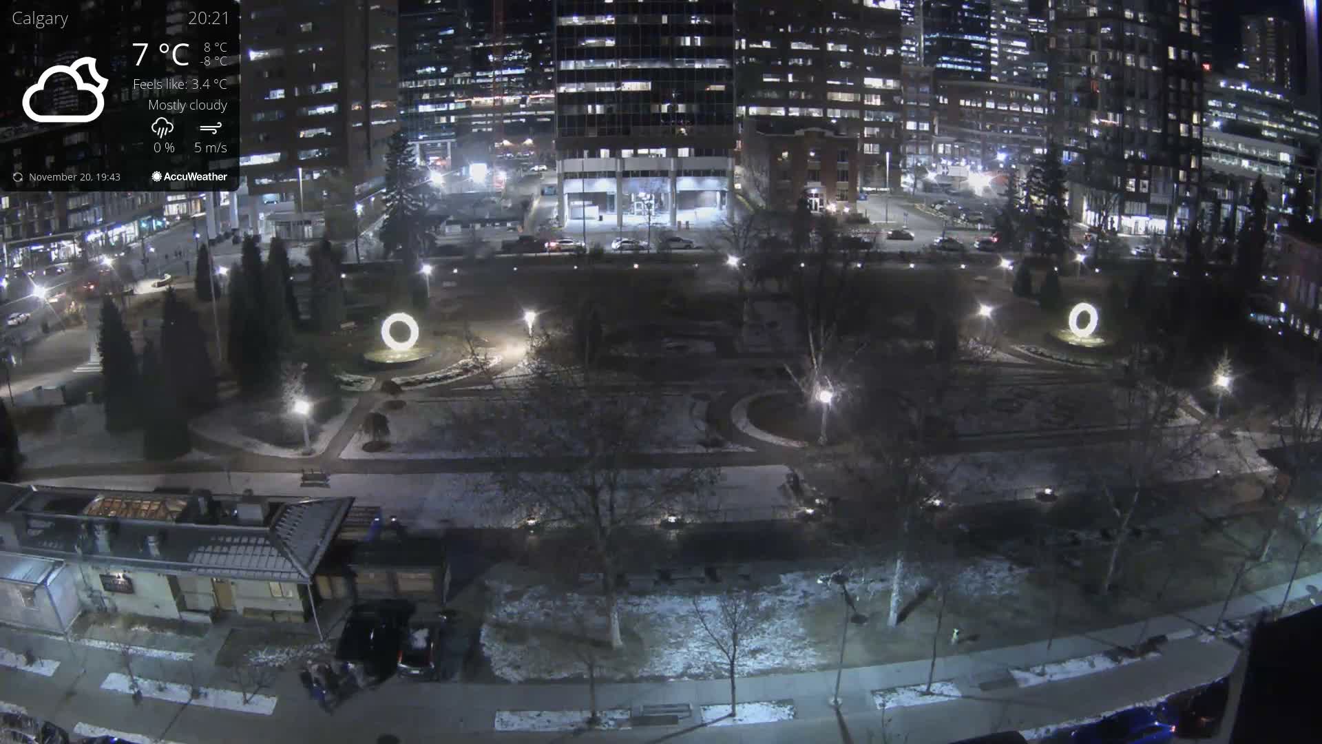 A nighttime, lightly raining view of a city park surrounded by illuminated buildings and trees.