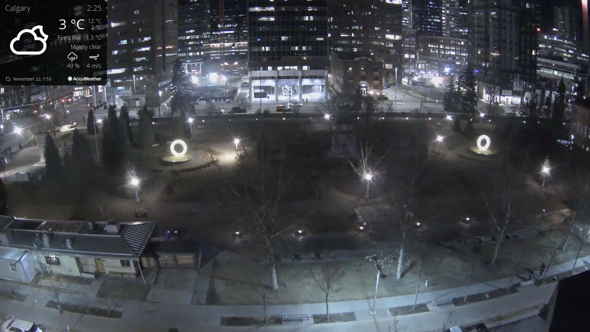 A nighttime aerial view captures a city park with bare trees, illuminated pathways, and glowing circular art installations, framed by brightly lit high-rise buildings and some residential structures, under a mostly clear sky.