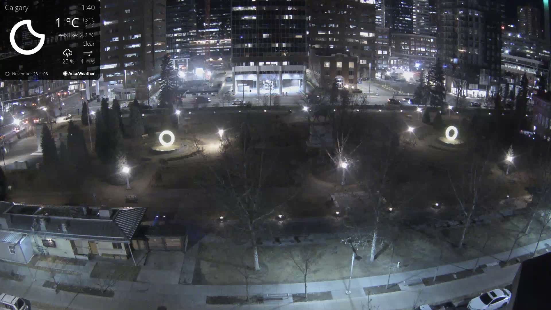 A wide overhead view captures a city park at night, illuminated by streetlights and featuring two glowing white ring sculptures among bare trees and paths, with a backdrop of brightly lit urban high-rises and busy streets under clear skies.