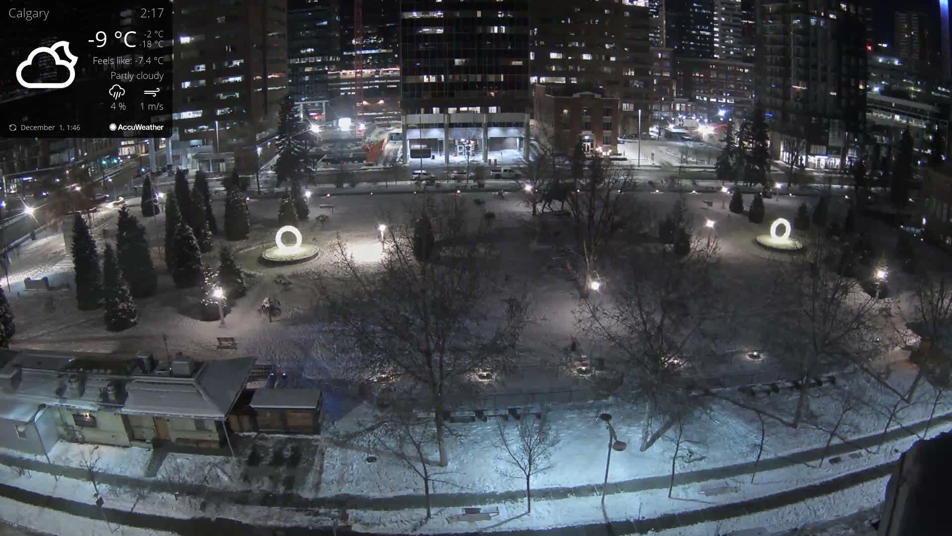 A snow-covered city park at night, illuminated by streetlights and a prominent glowing circular art installation, is viewed from above with tall urban buildings in the background, under cold winter conditions.