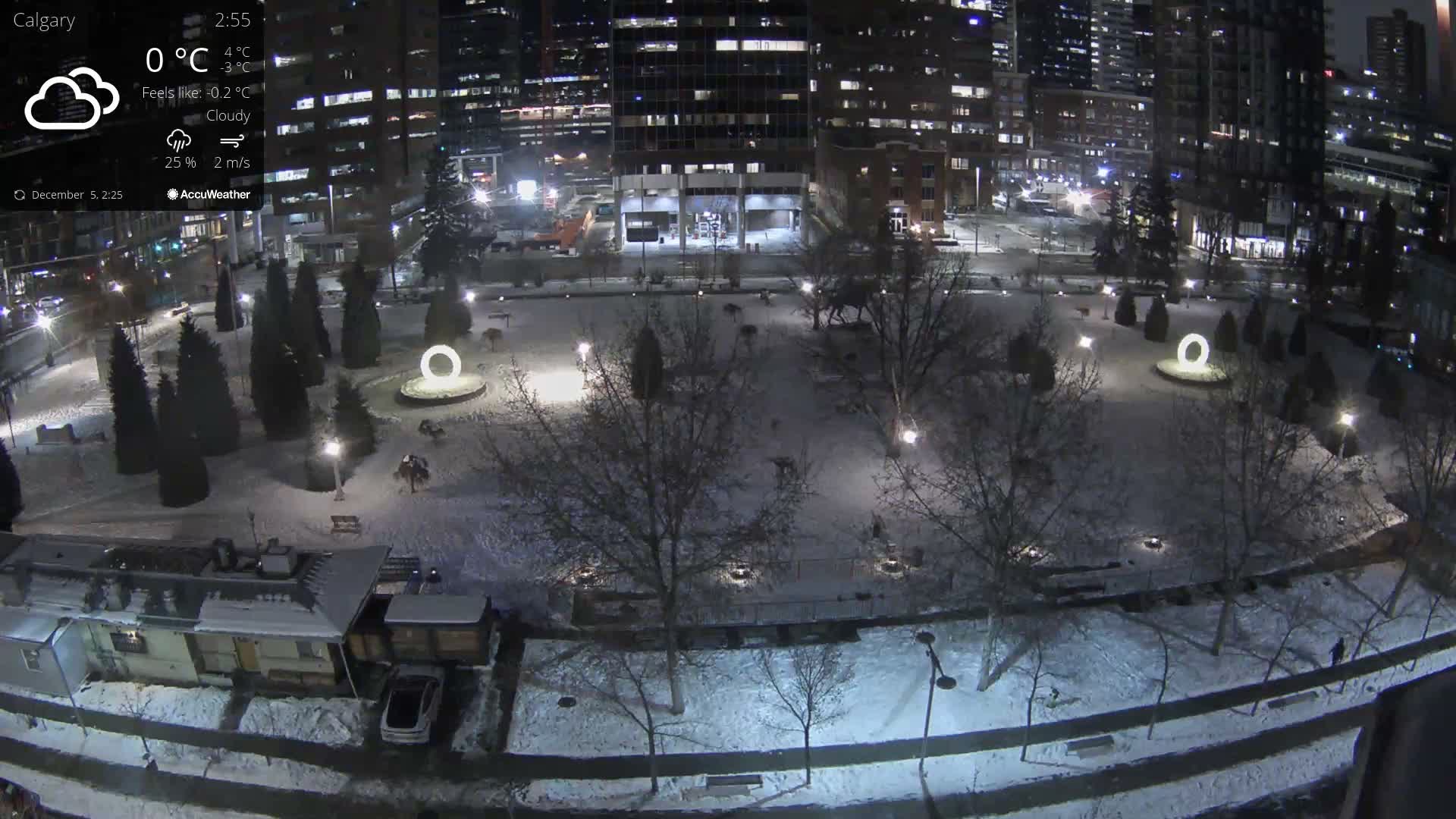 A snow-covered urban park at night, featuring illuminated paths, trees, and two large glowing circular structures, is seen from above, surrounded by brightly lit city buildings and roads under a cold, dark winter sky.