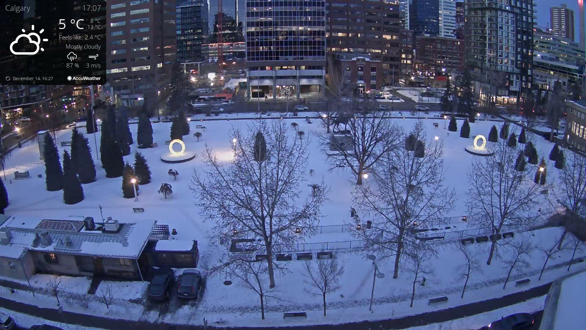 A snow-covered urban park at night, featuring illuminated paths, trees, and two large glowing circular structures, is seen from above, surrounded by brightly lit city buildings and roads under a cold, dark winter sky.