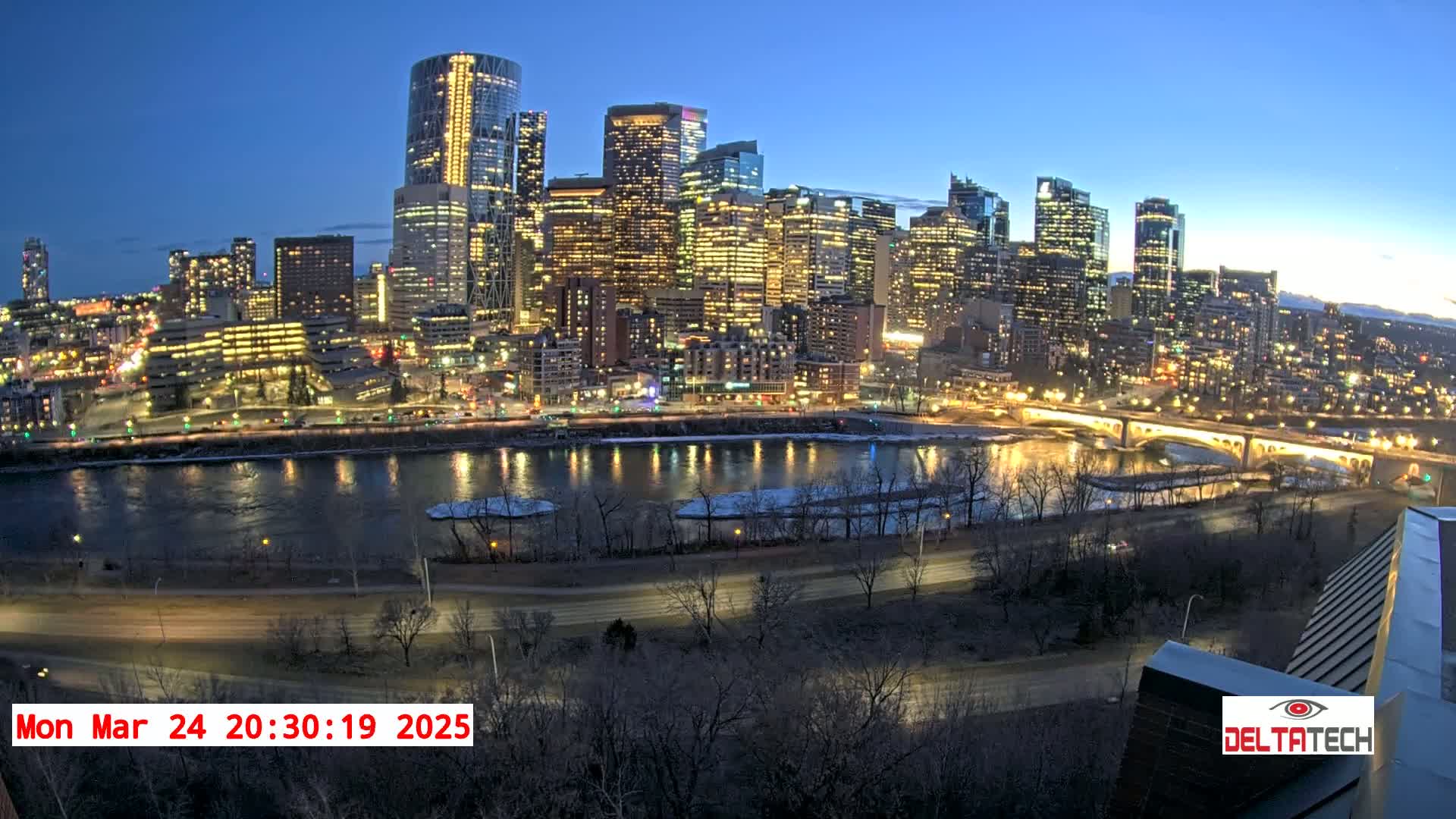 A brightly lit city skyline at twilight is situated across a river with a partially frozen shoreline.