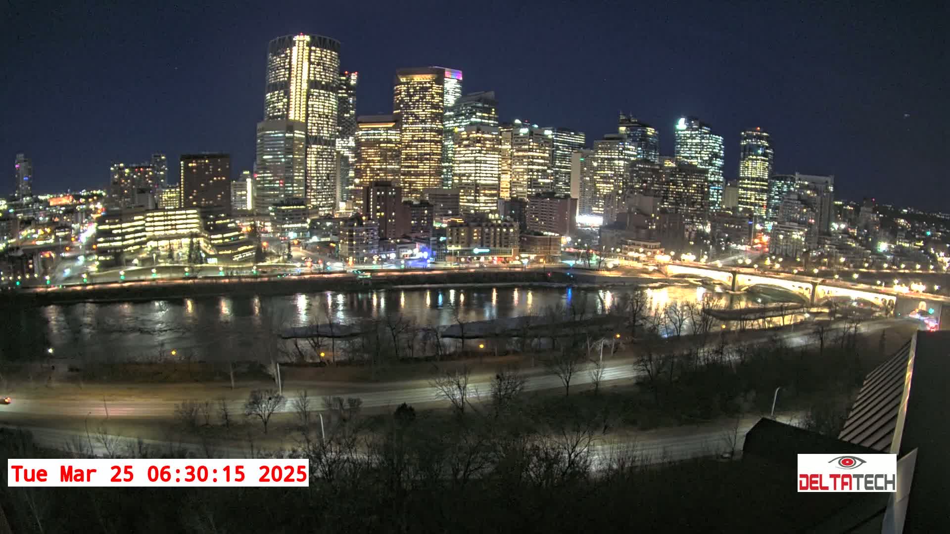 A nighttime cityscape with brightly lit skyscrapers sits across a river from a roadway, under a clear, dark sky.