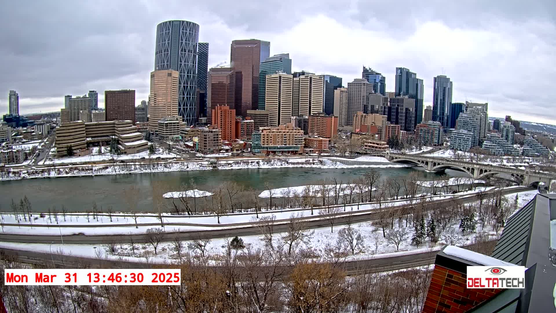 A snow-covered cityscape with a river and bridge under a cloudy sky.
