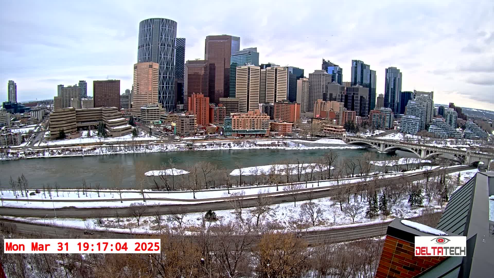 A snow-covered city skyline with numerous skyscrapers is seen across a river with a bridge, under a cloudy sky.