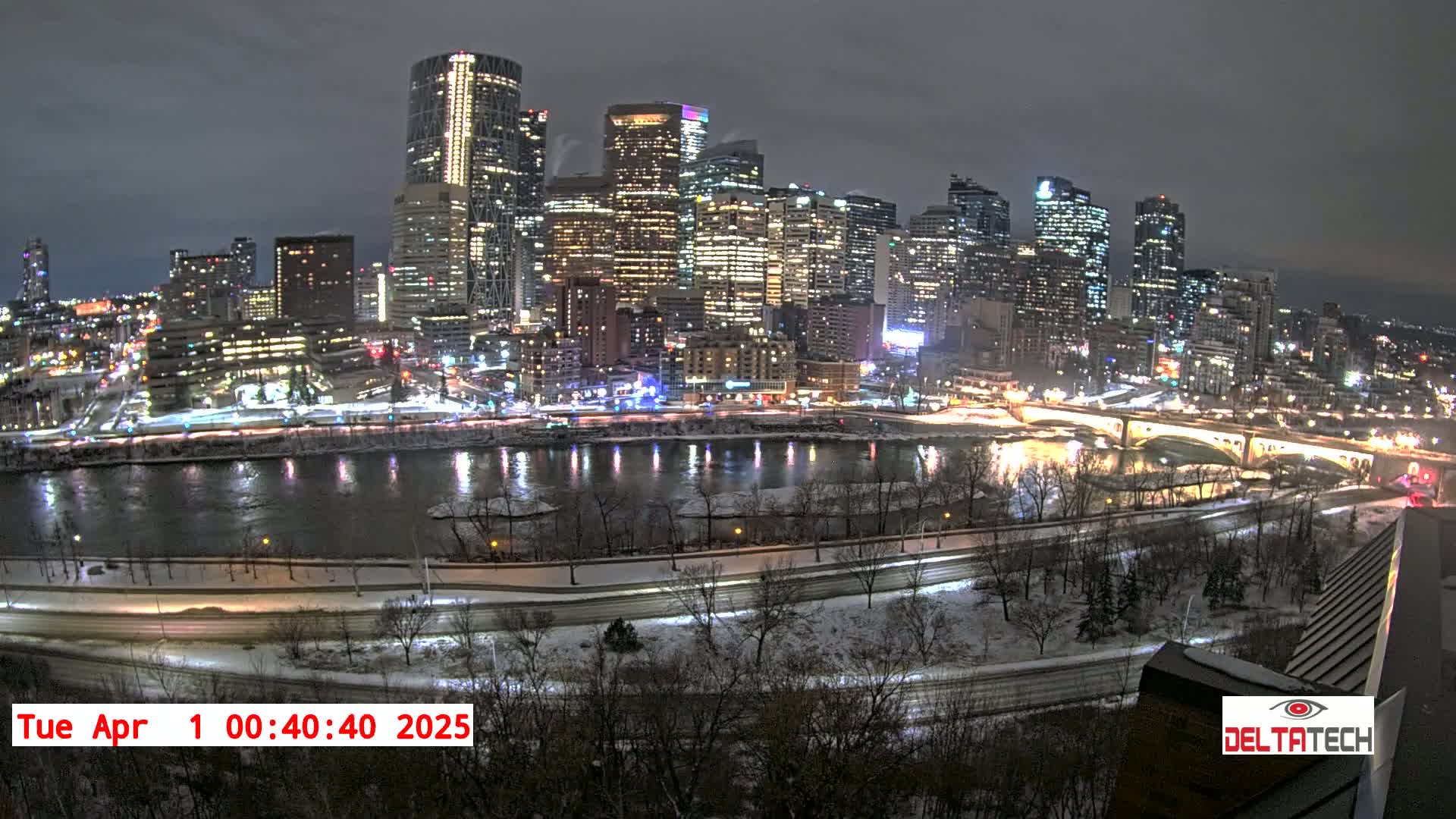 A snow-covered city skyline at night is illuminated by bright lights reflecting on a river, with a bridge visible in the distance.