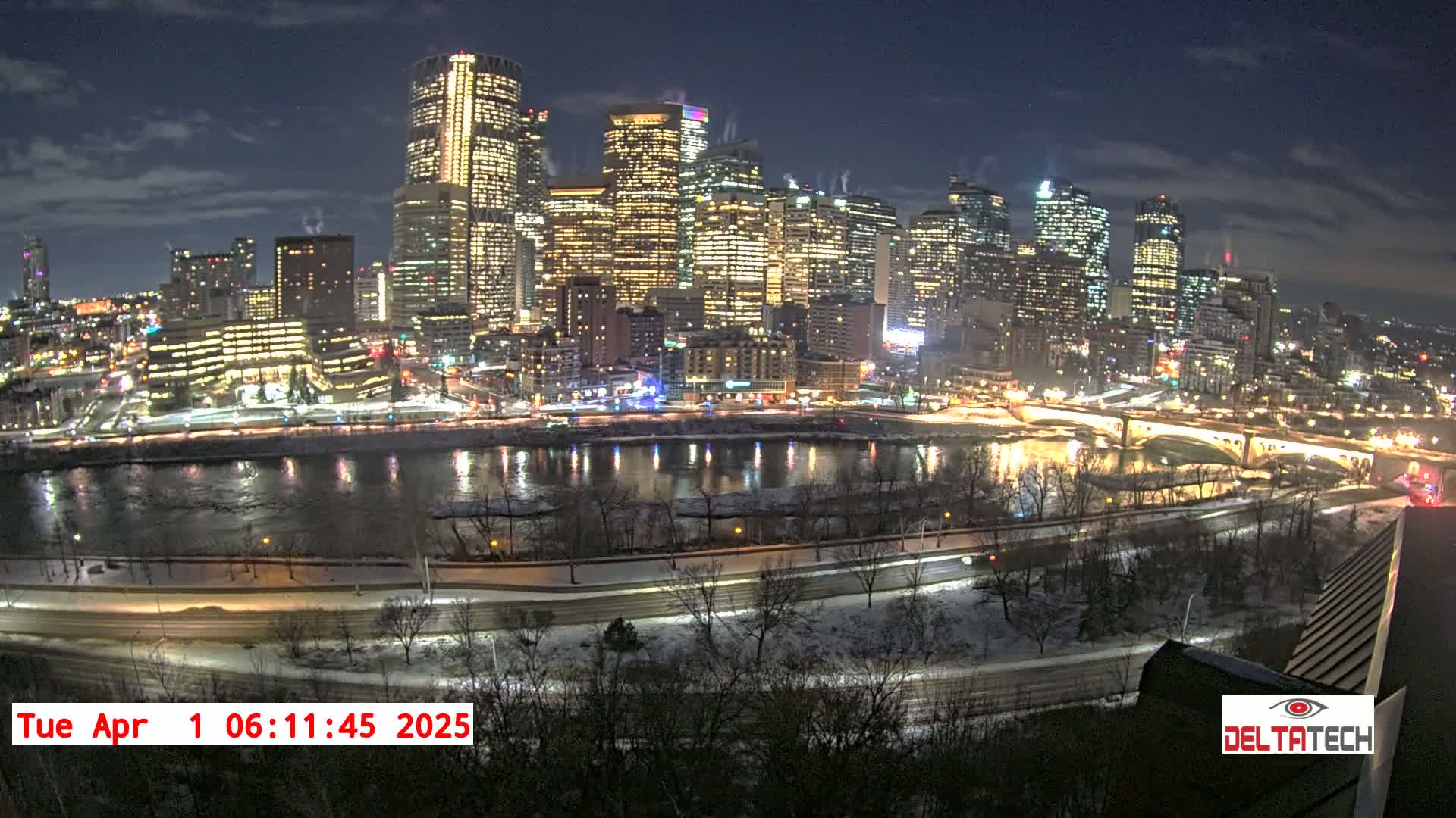 A nighttime cityscape with illuminated skyscrapers and bridges along a snow-dusted riverbank under a partly cloudy sky.
