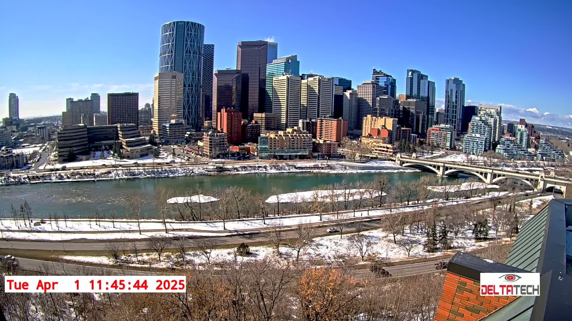 A sunny cityscape with snow-covered ground and a river partially frozen, viewed from an elevated perspective.
