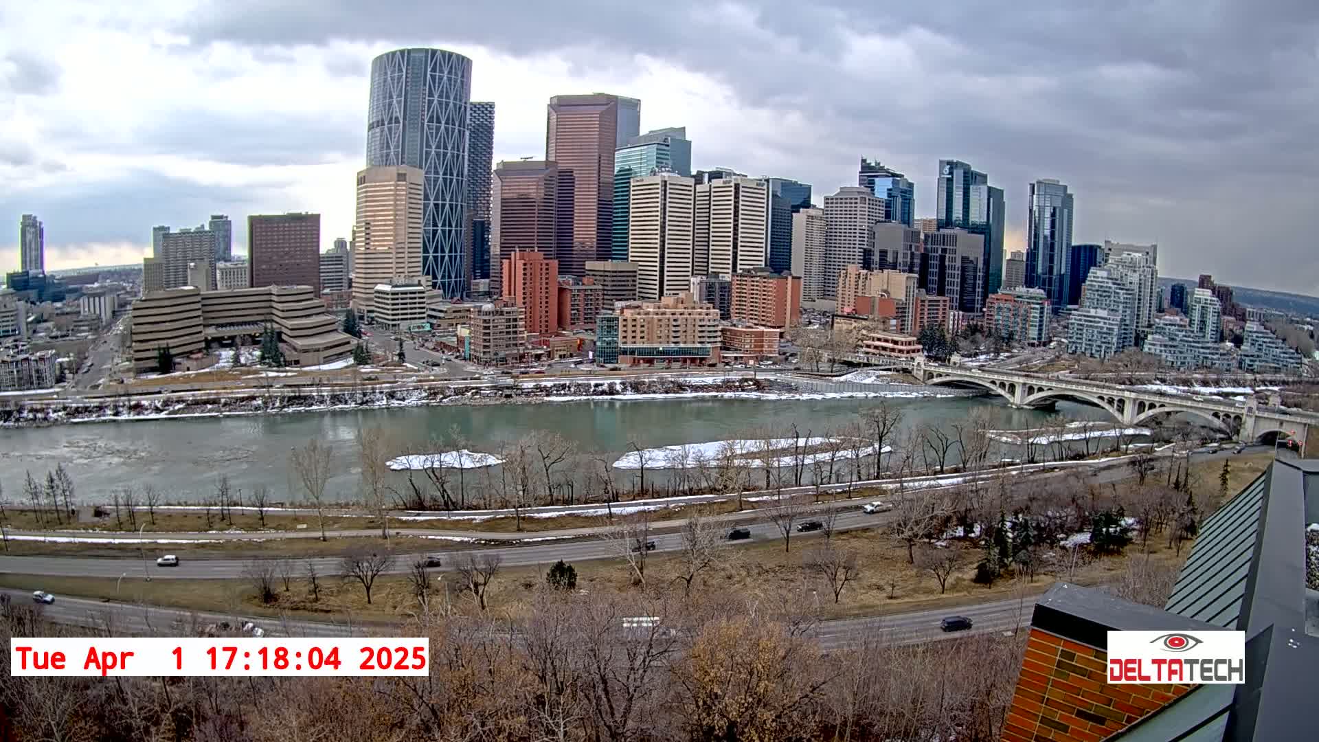 A city skyline with numerous skyscrapers is visible across a river with patches of ice and snow under a cloudy sky.